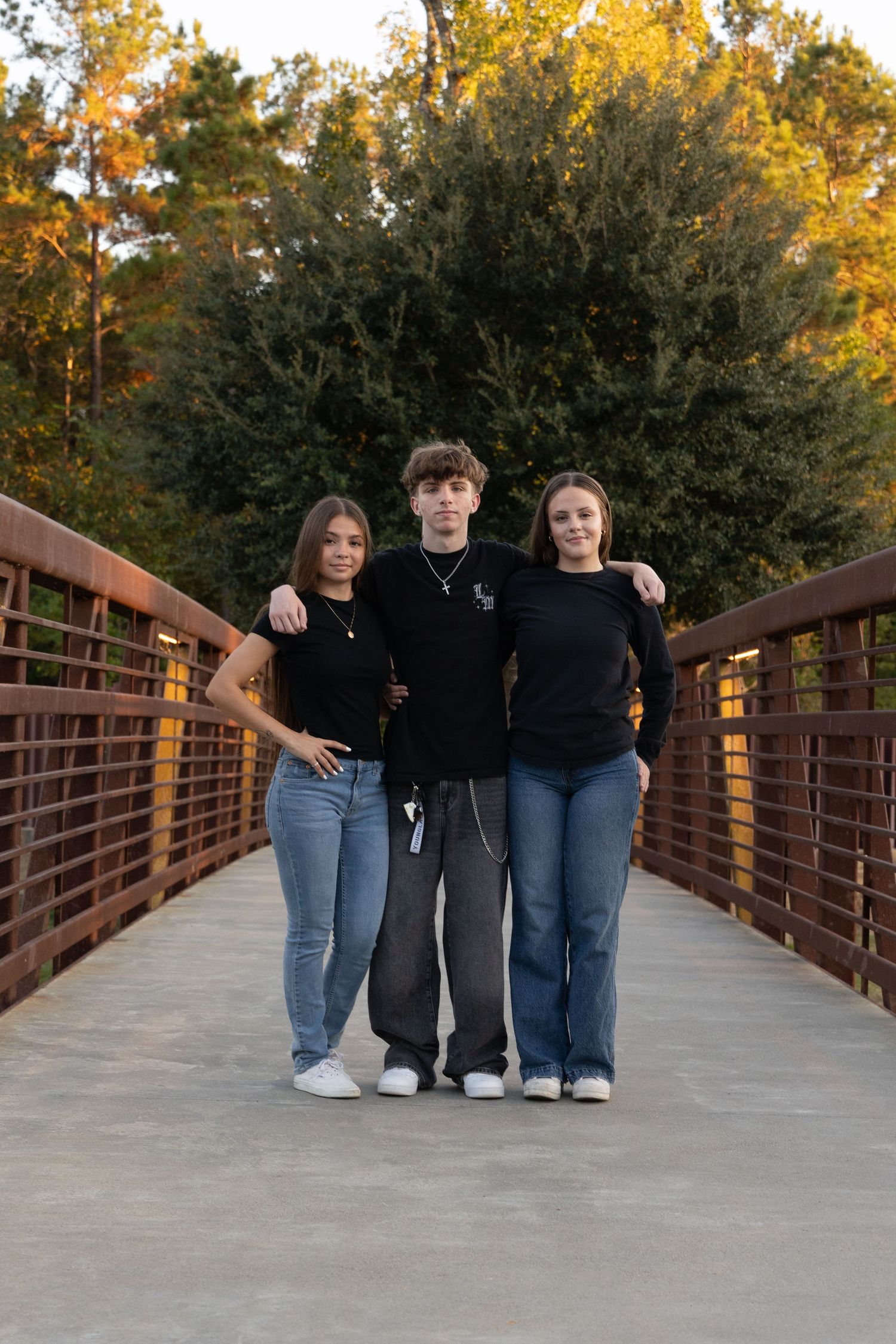 Three teens pose on a wooden bridge surrounded by autumn trees at golden hour, wearing black tops and jeans.