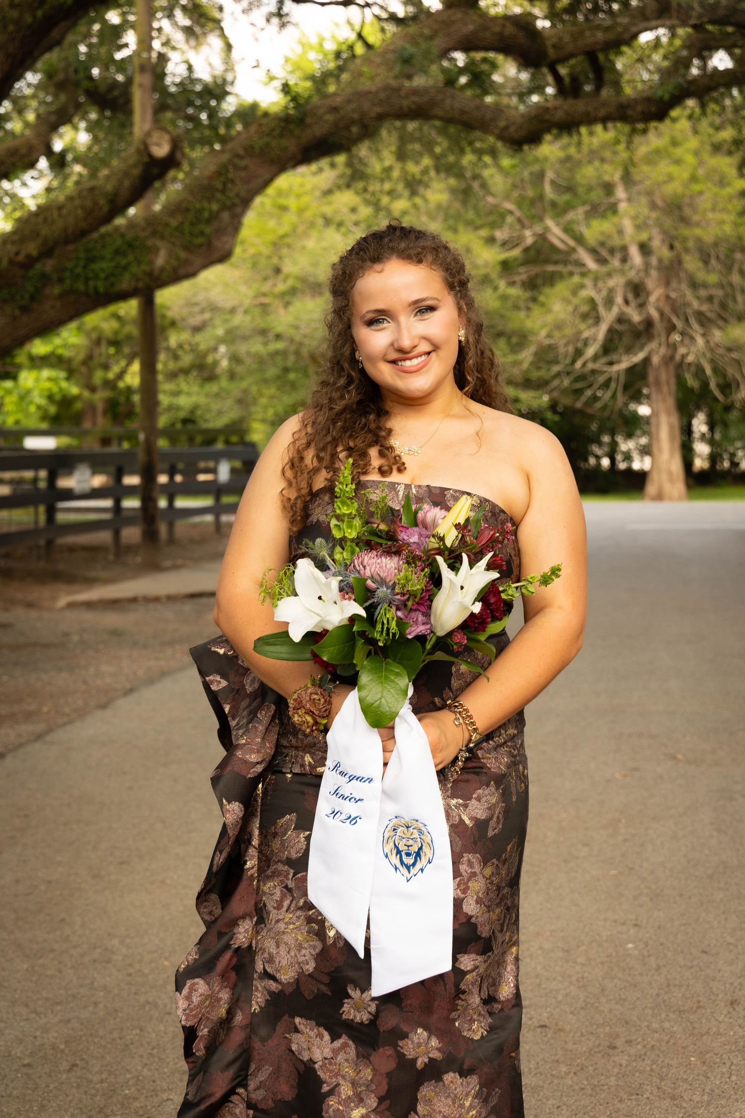 Young woman in floral strapless gown holding bouquet with white sash outdoors under oak trees.