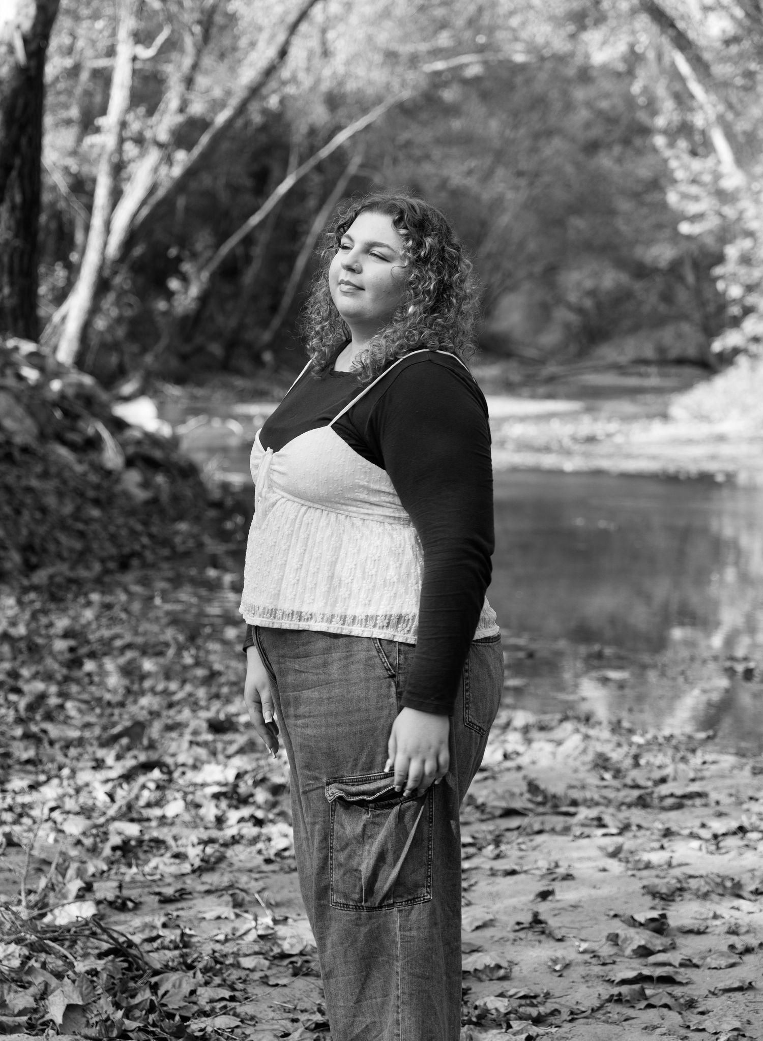 Black and white portrait of a curly-haired woman standing outdoors near a creek, wearing a cami top and long skirt.