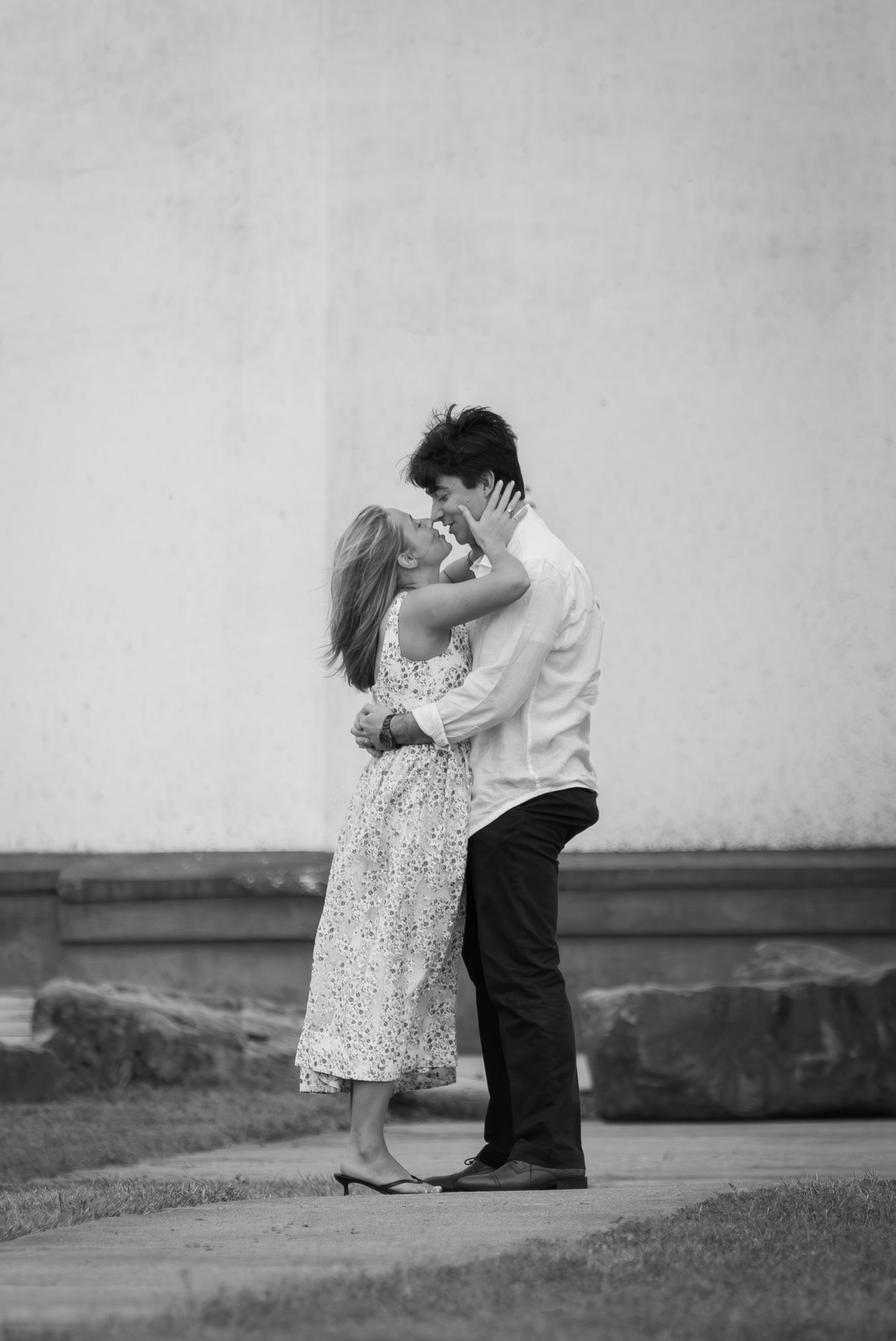 Black and white photo of romantic couple embracing and kissing outdoors against a light wall.