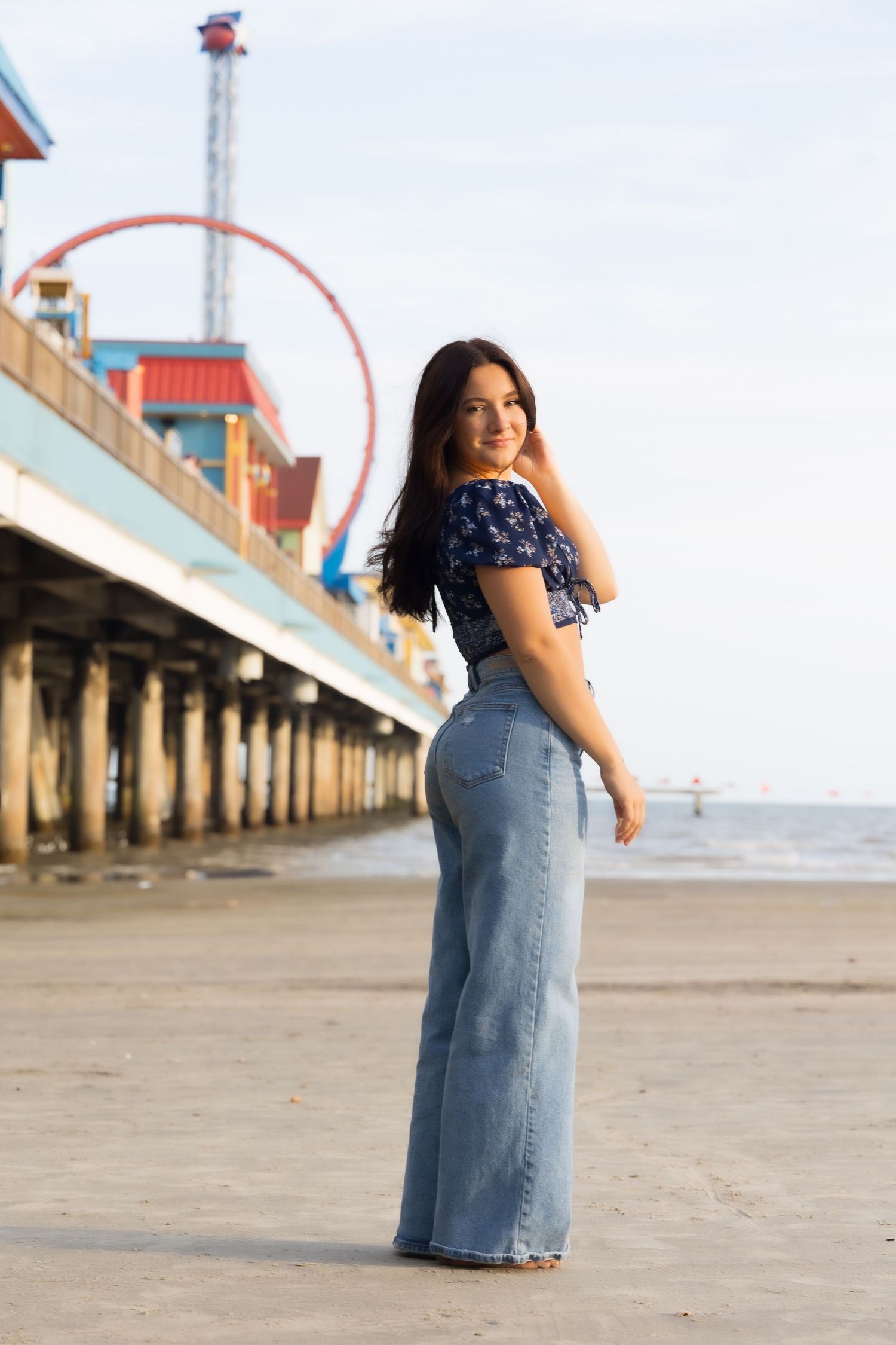 Young woman in wide-leg jeans and floral top poses on beach near amusement pier with Ferris wheel backdrop.
