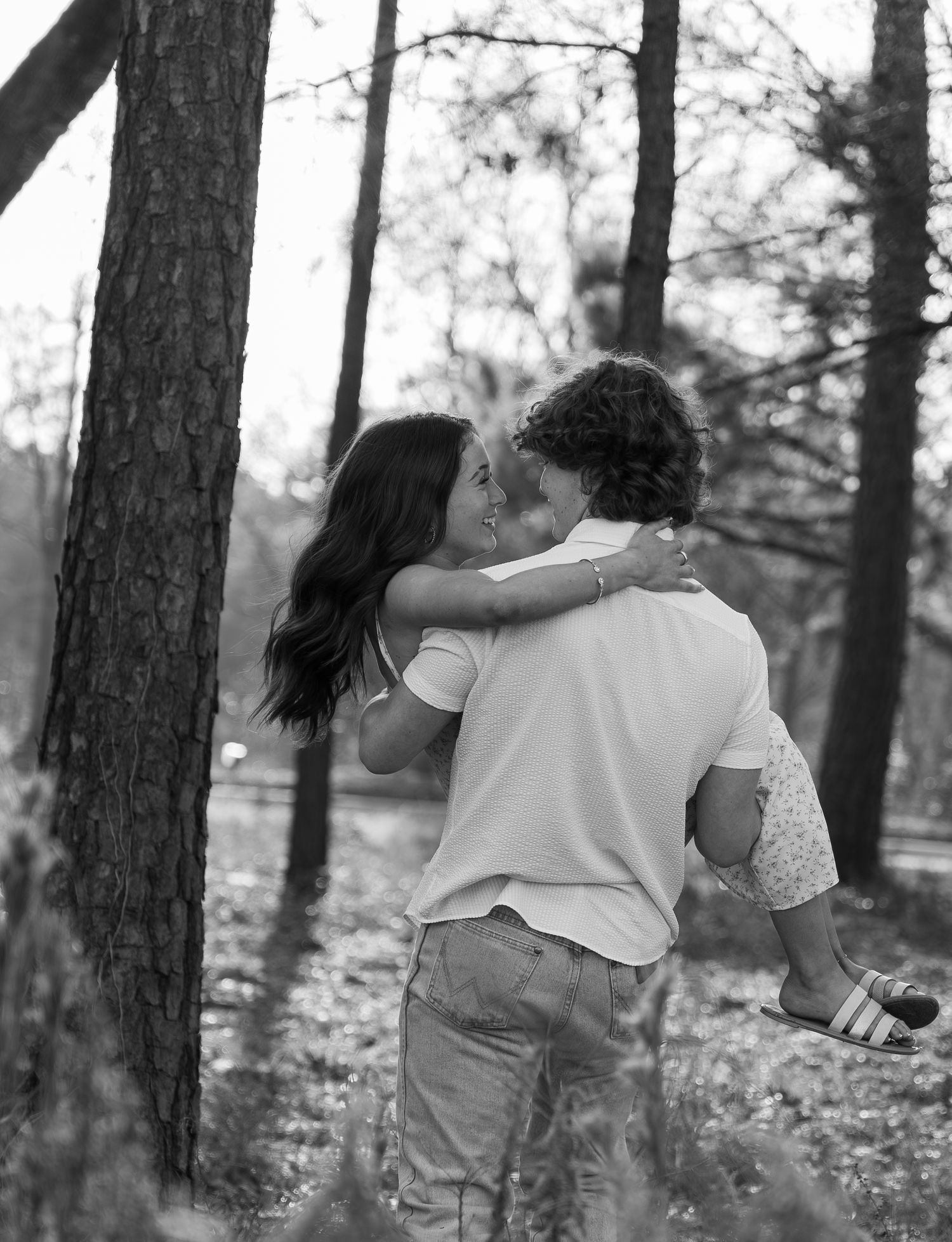 Black and white photo of a couple sharing a romantic kiss in a forest, man lifting woman among tall trees.