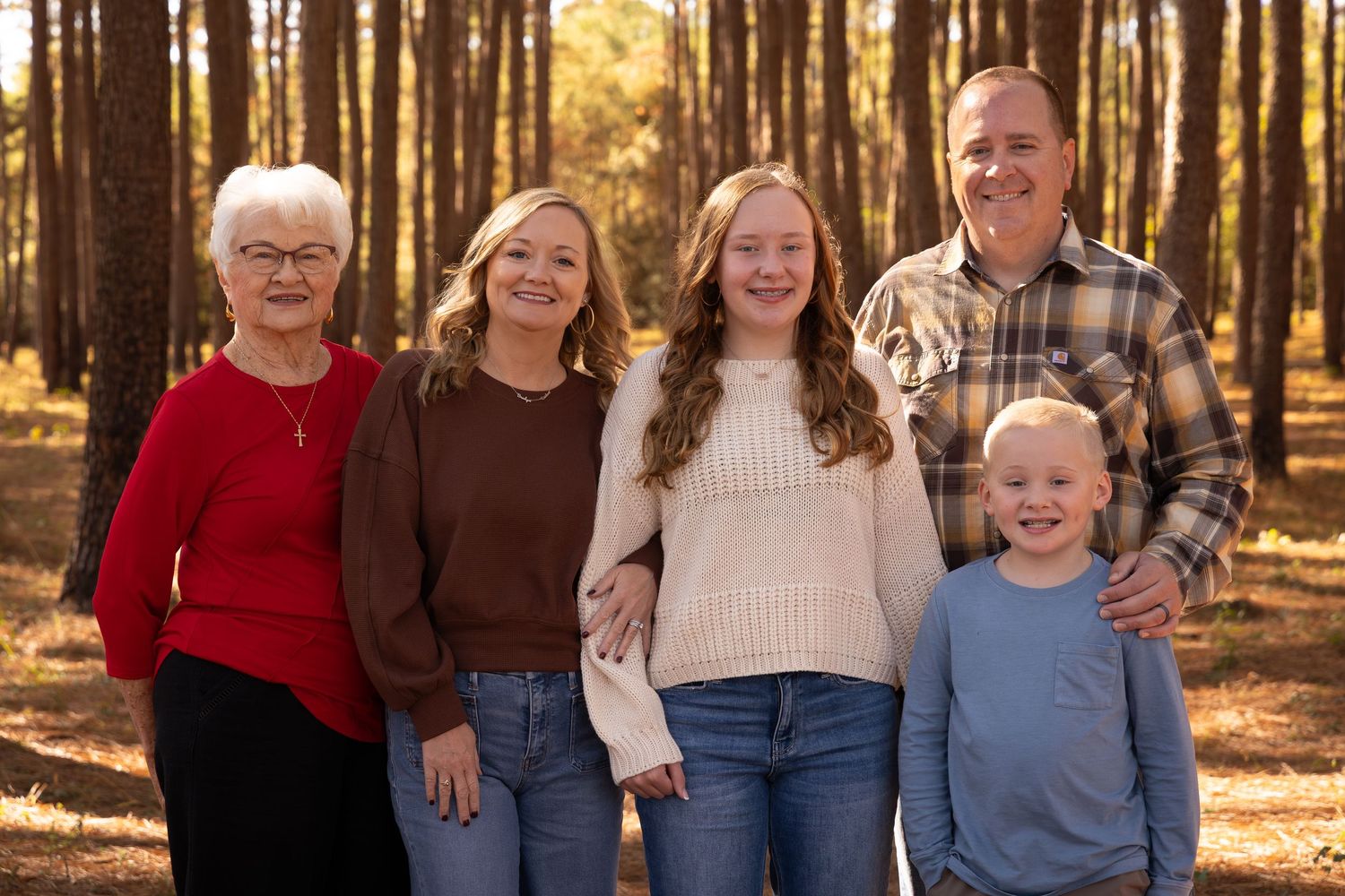 Multi-generational family of five smiling together among tall pine trees in a warm autumn forest setting.