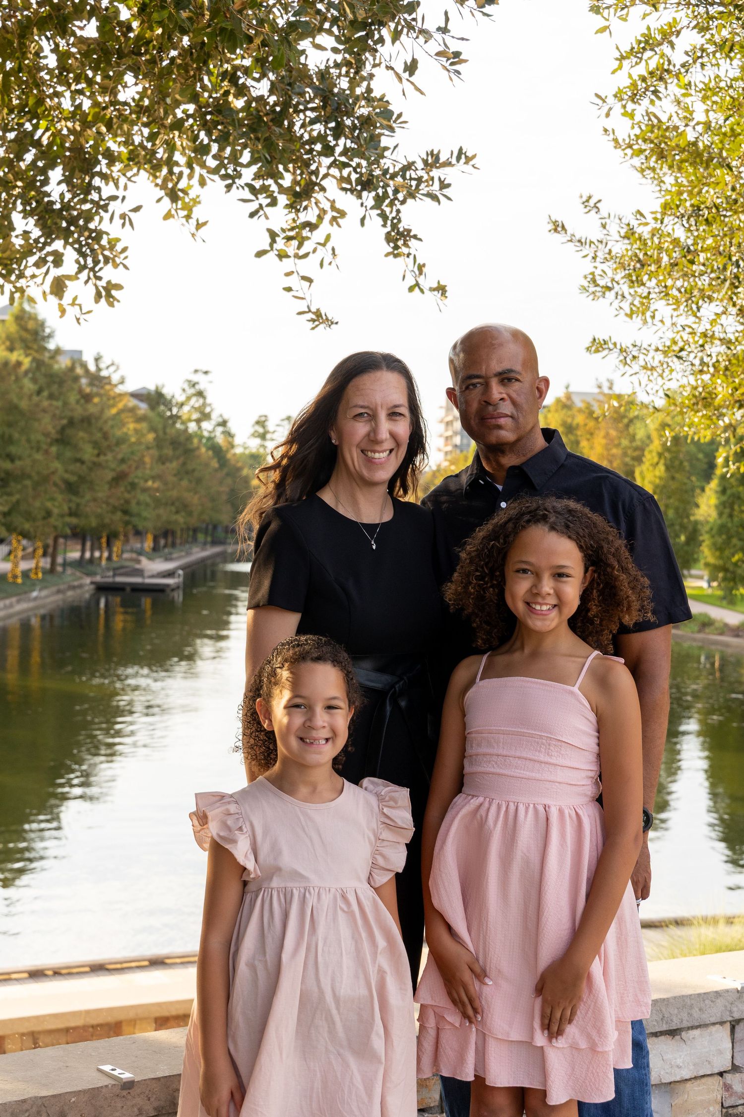 Family of four smiling outdoors near a scenic waterway, with two girls in pink dresses and parents standing behind them.