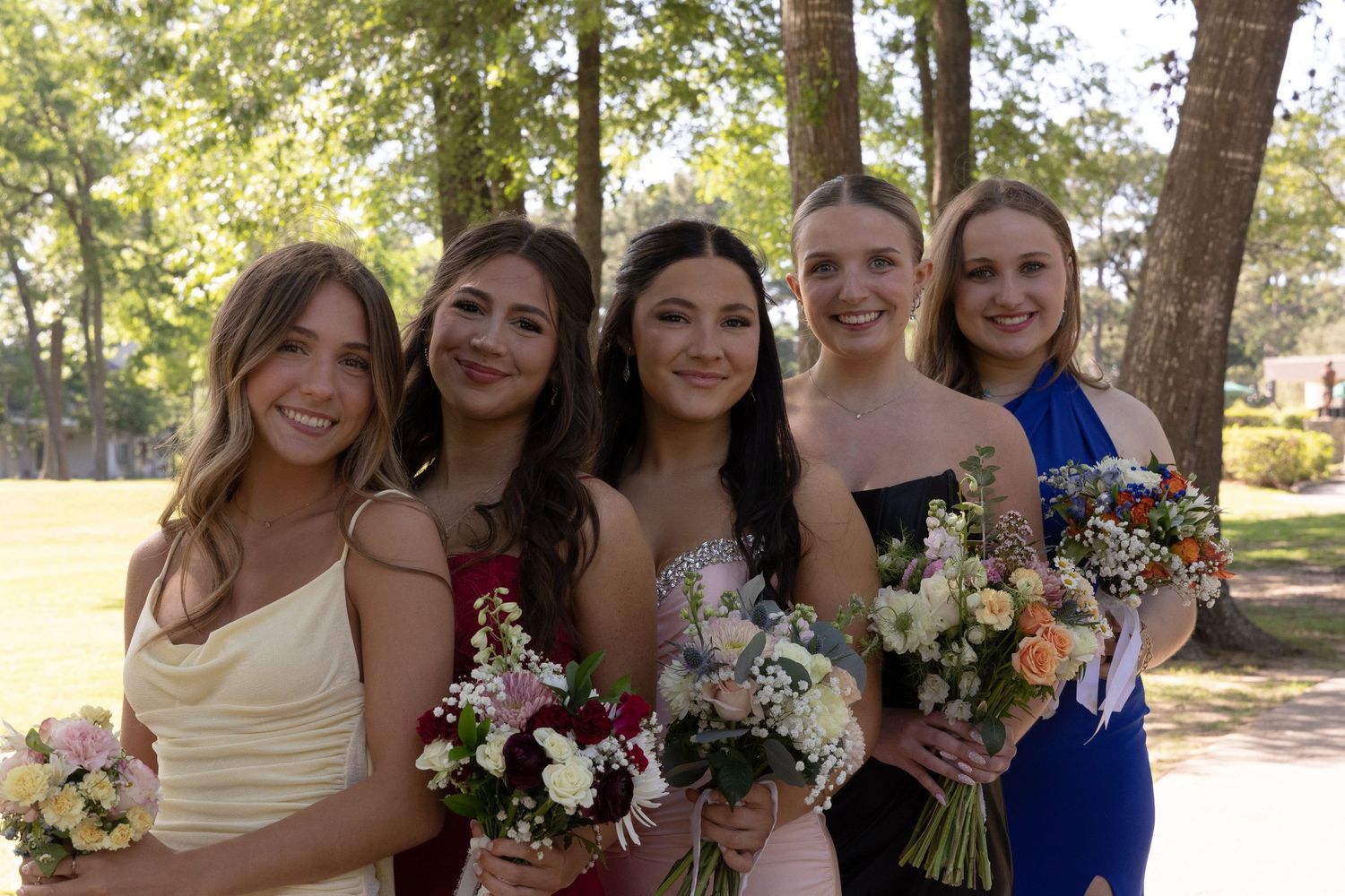 Five young women in formal dresses holding bouquets of flowers pose together outdoors at prom.
