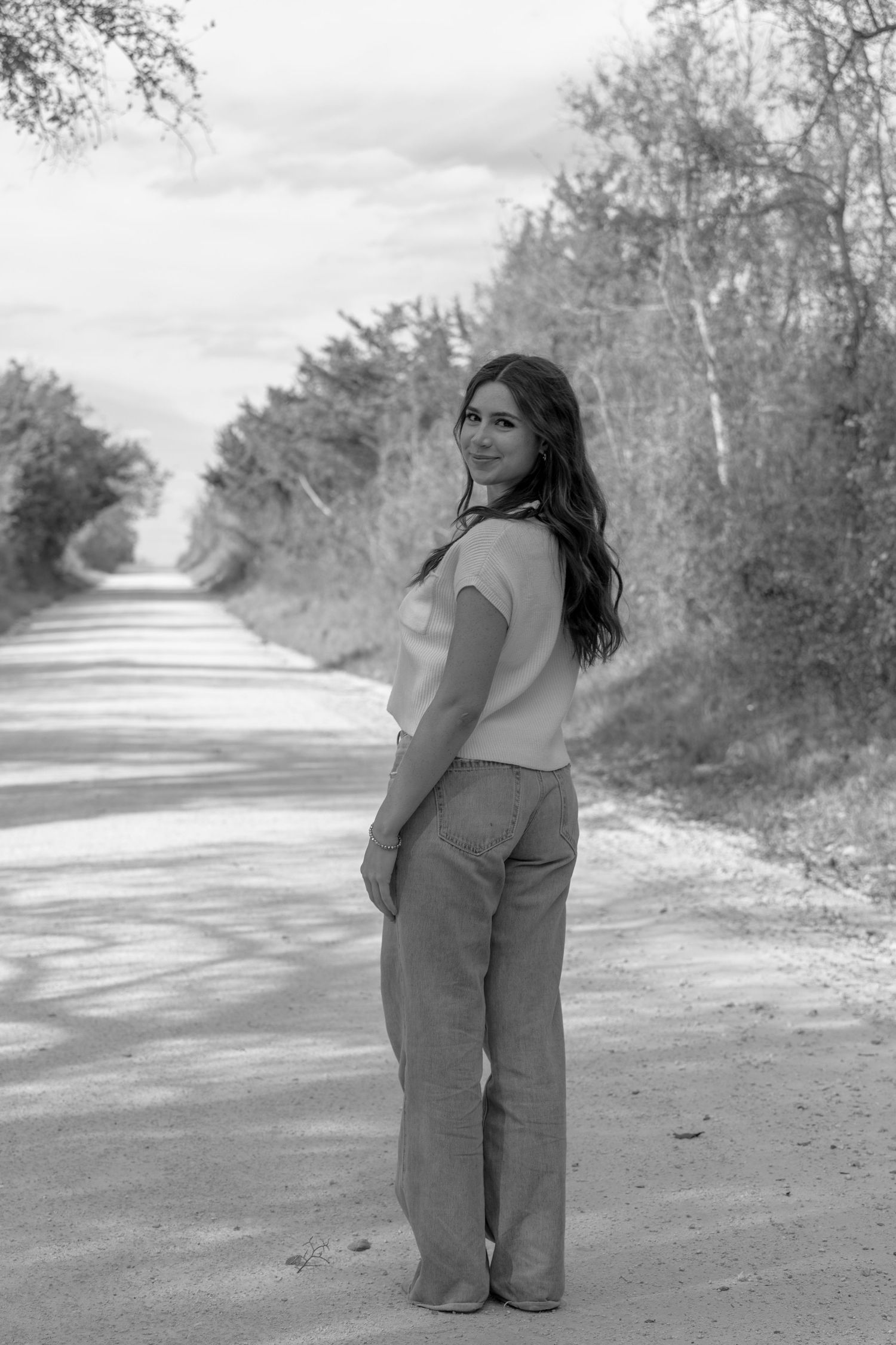Black and white portrait of a person standing on a rural dirt road surrounded by trees.