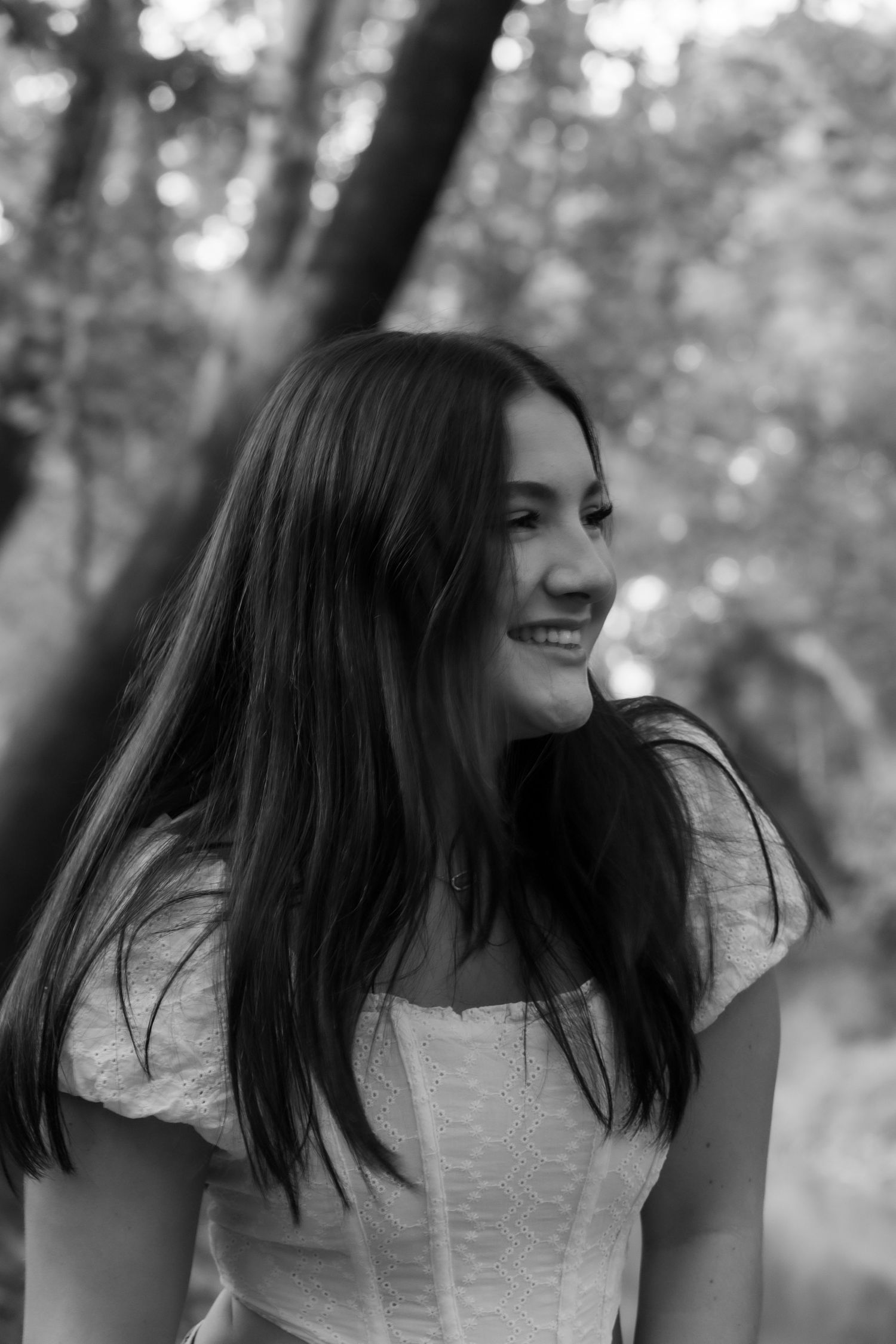 Black and white portrait of a smiling young woman with long hair in a white dress, posing outdoors among trees.