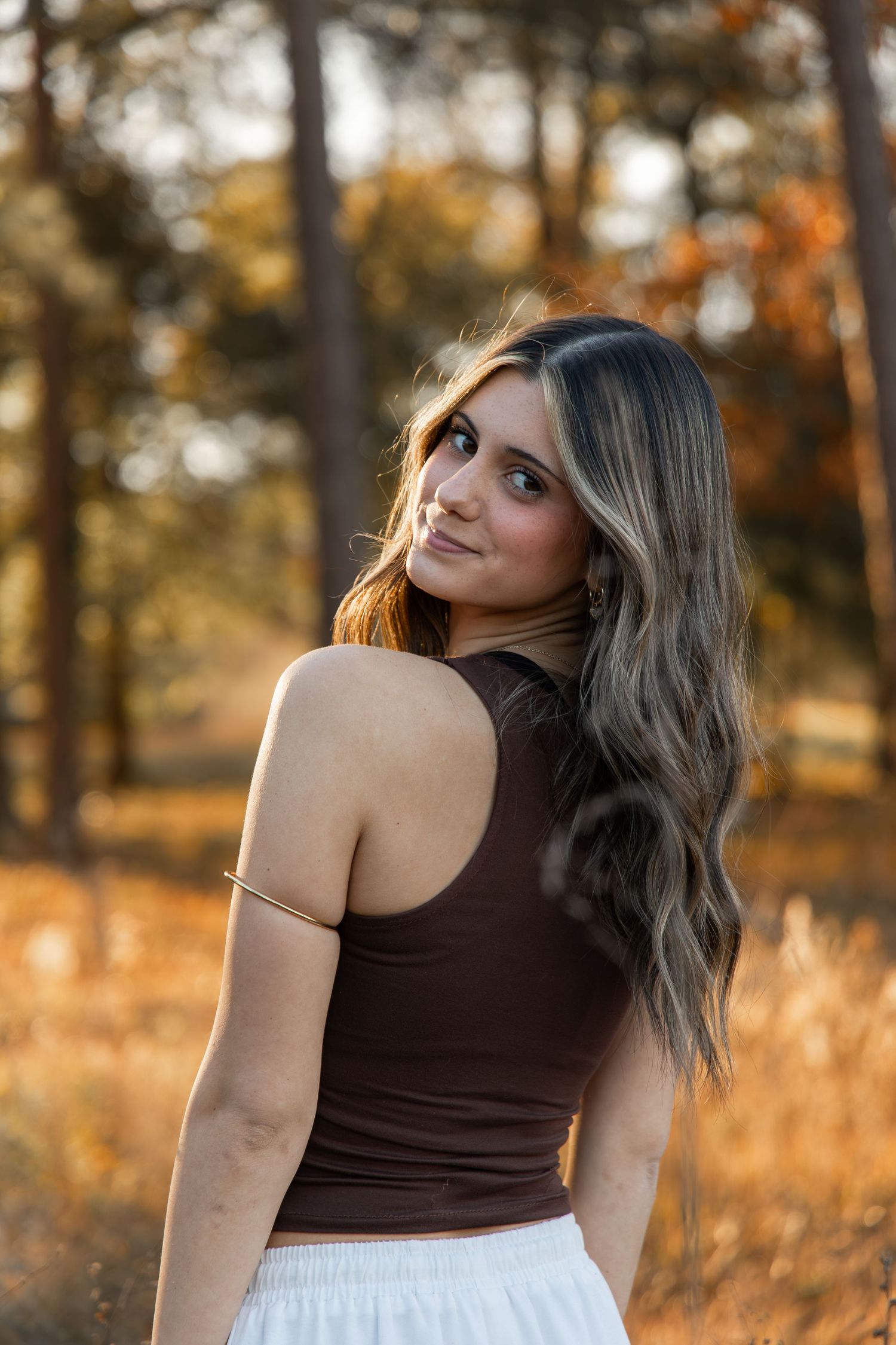 Young woman with long wavy hair in brown crop top and white skirt glancing over shoulder in autumn forest setting.