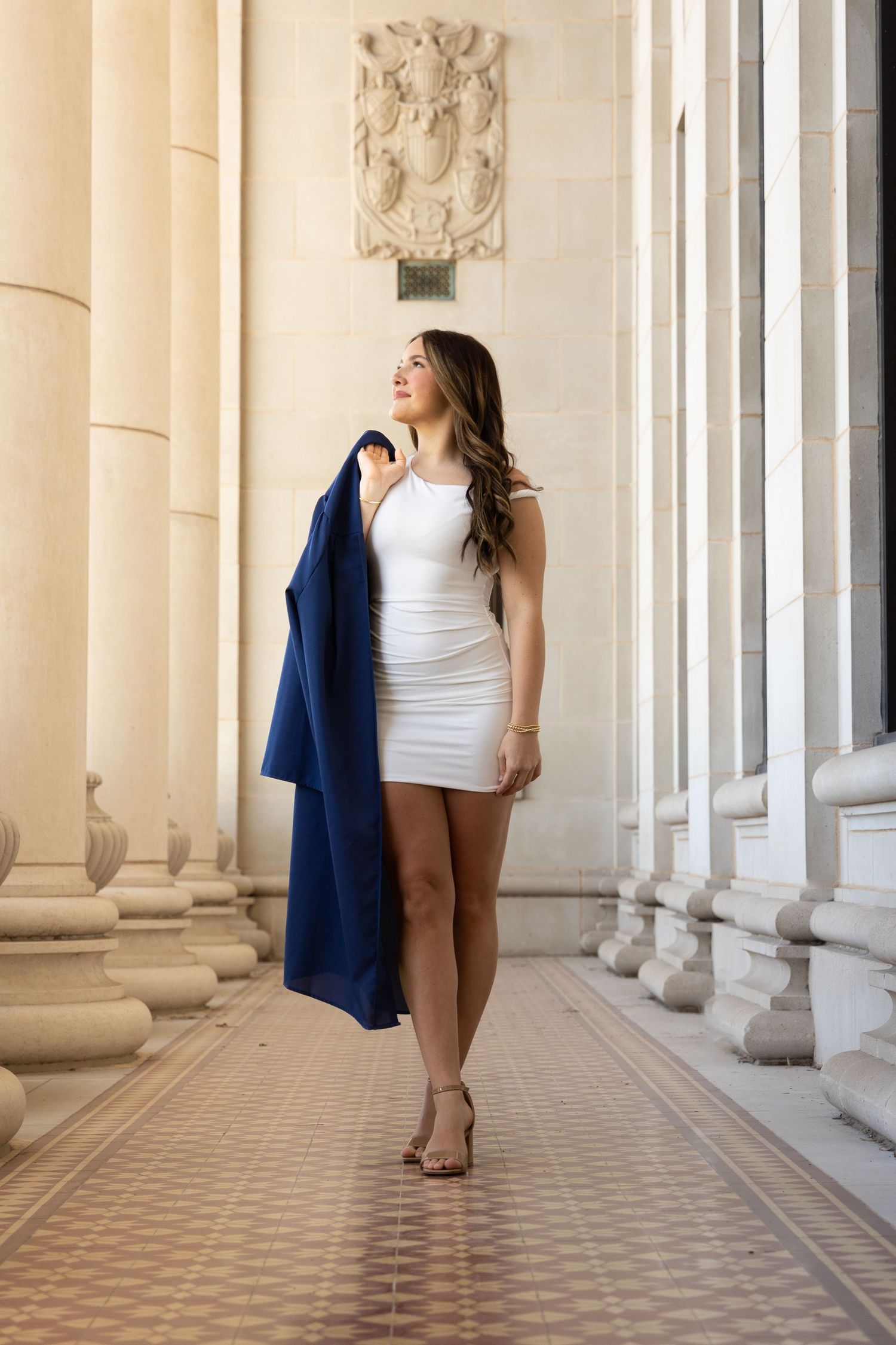Young woman in white mini dress holding navy blazer, posing among stone columns of elegant building.