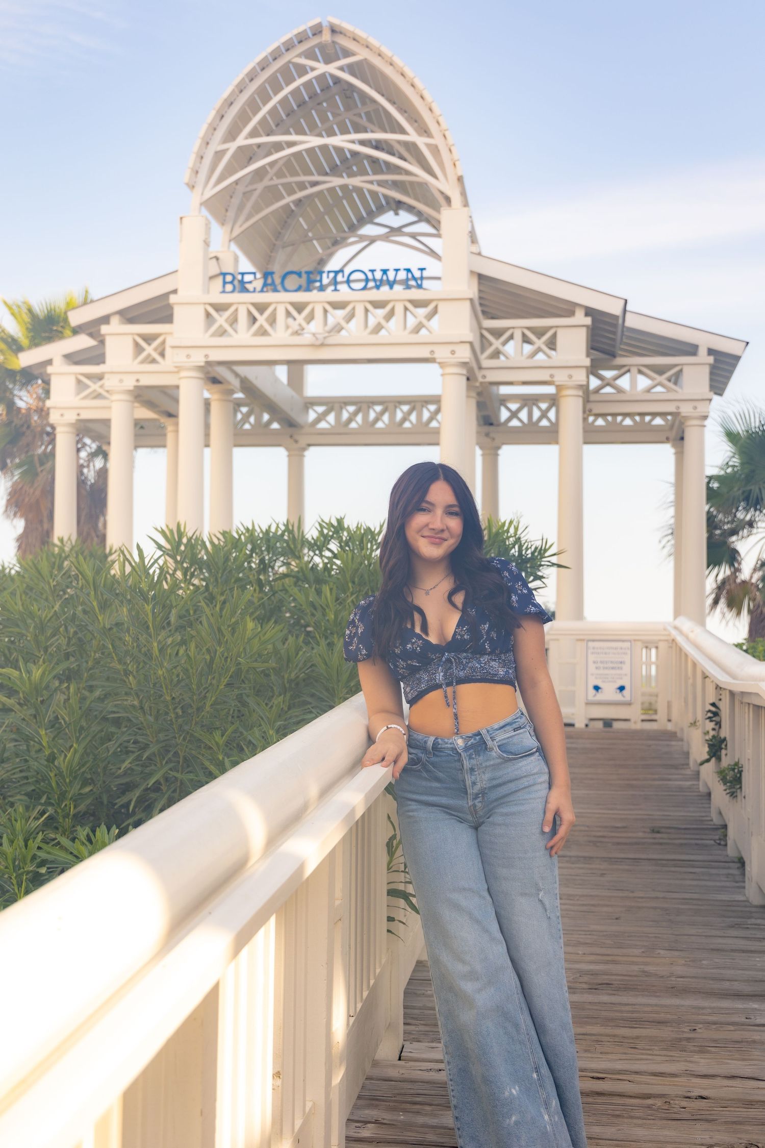 Young woman in crop top and jeans posing on boardwalk in front of Beachtown sign and white pavilion.