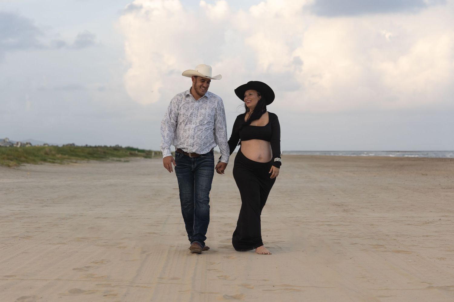 Pregnant couple holding hands walking on a sandy beach, man in cowboy hat and woman in black maternity dress.