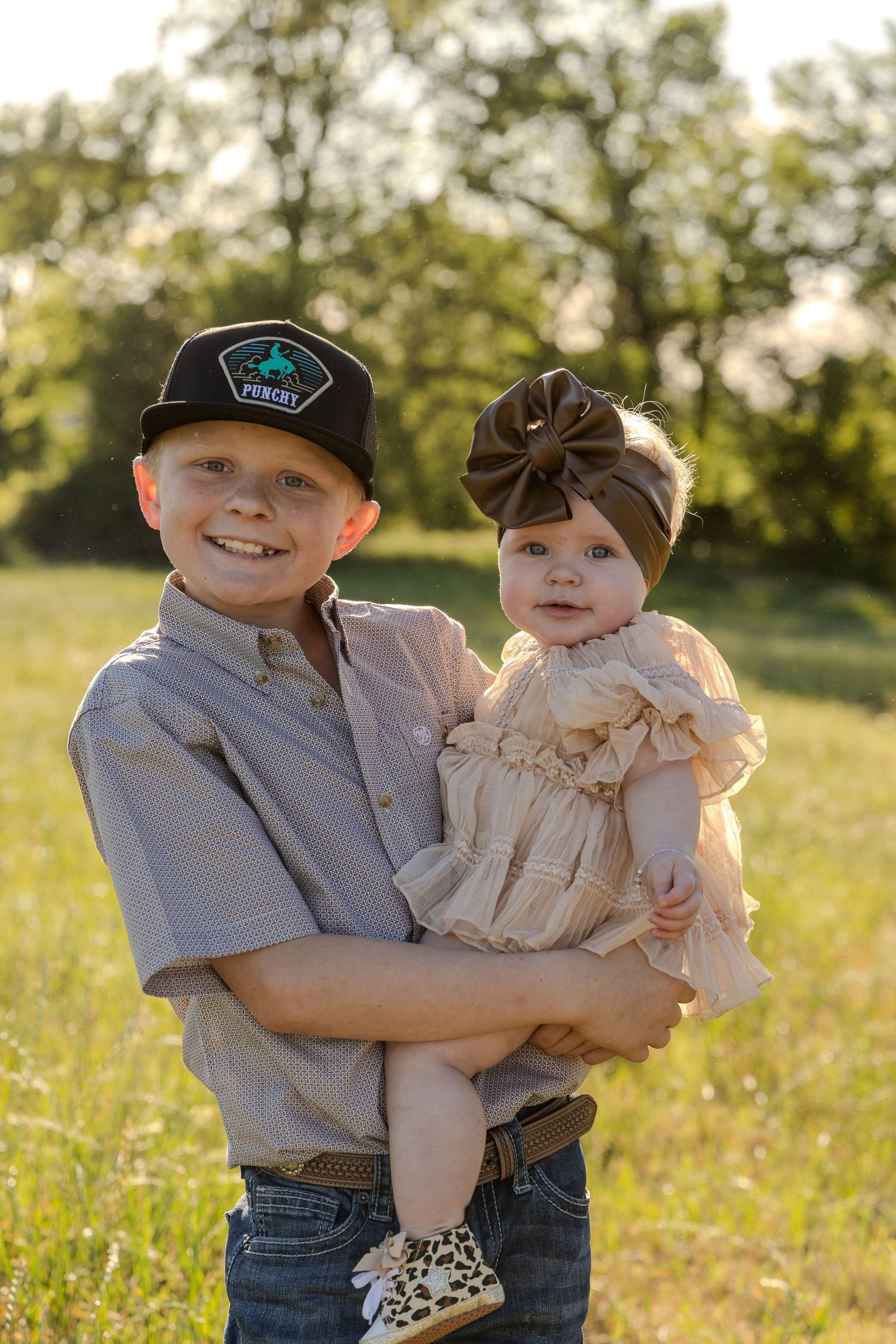 A smiling boy in a cap holds a baby girl in a ruffled dress outdoors in a sunny green field.