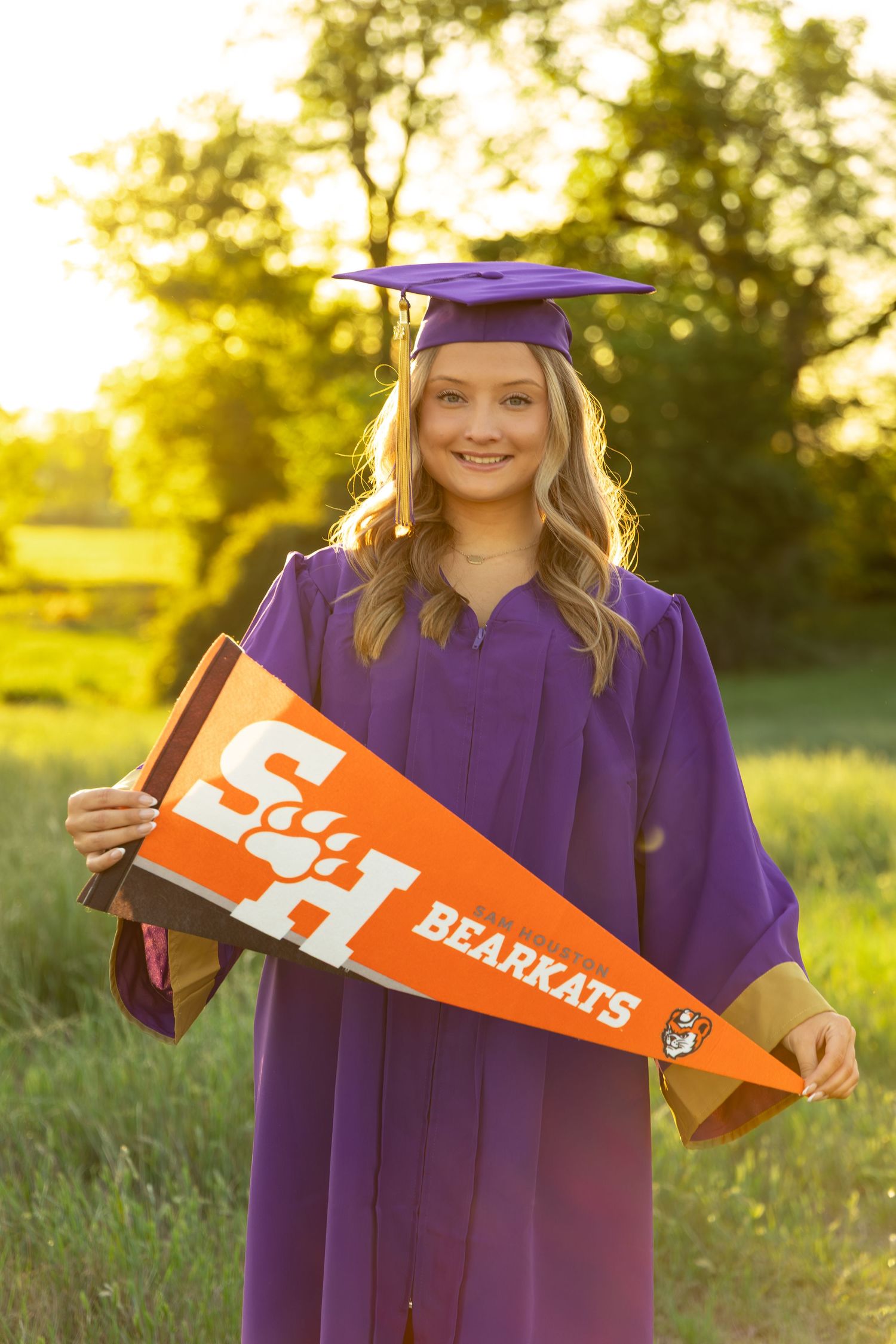 Smiling graduate in purple cap and gown holding Sam Houston State Bearkats orange pennant outdoors in golden sunlight.