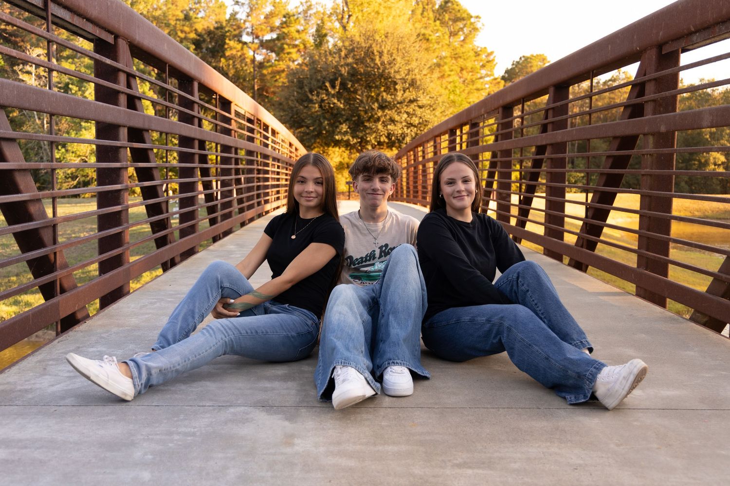Three teenagers sitting together on a wooden bridge outdoors, smiling, wearing jeans and dark tops during golden hour.