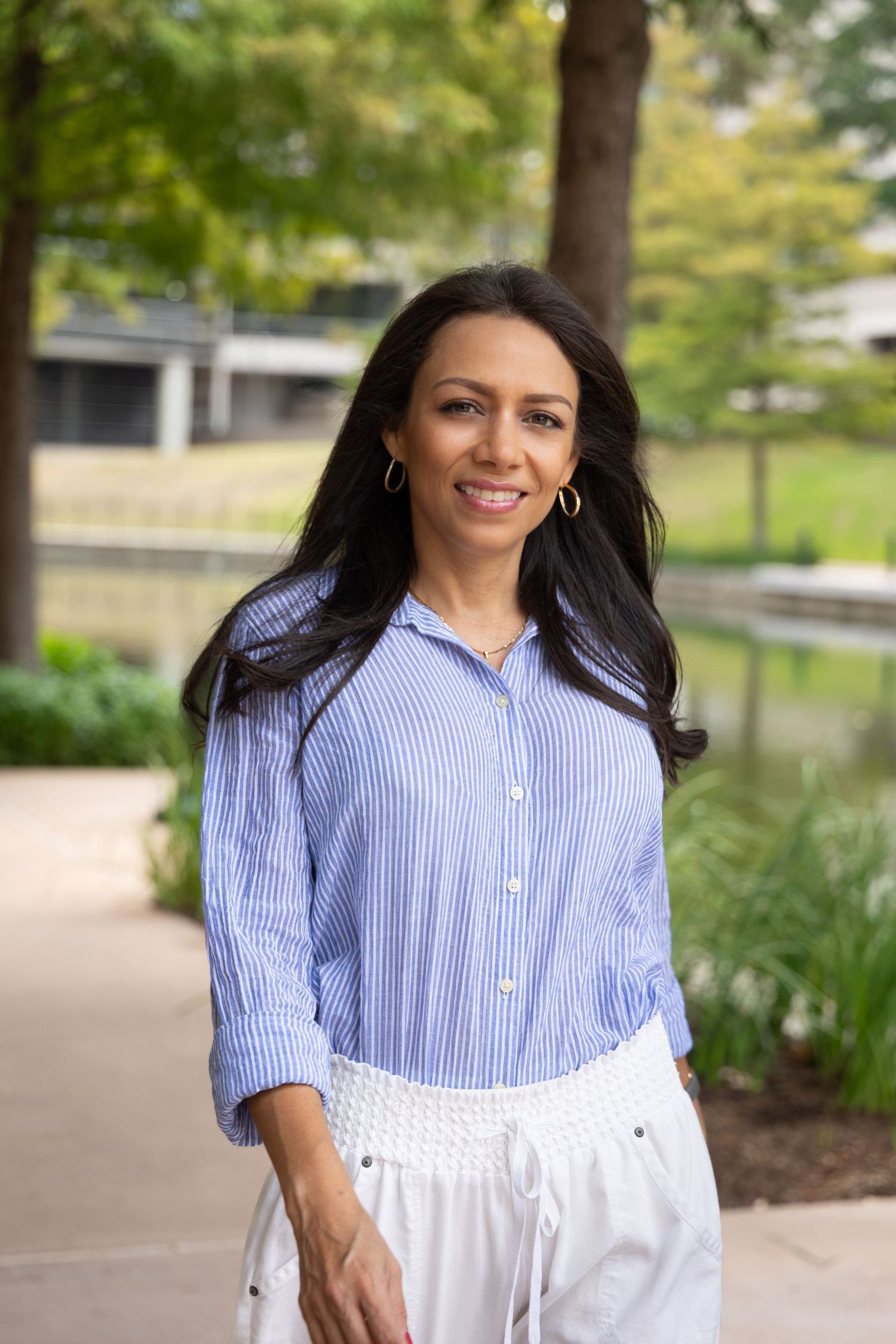 Smiling woman in blue striped shirt and white pants poses outdoors near a reflective pond with green trees.