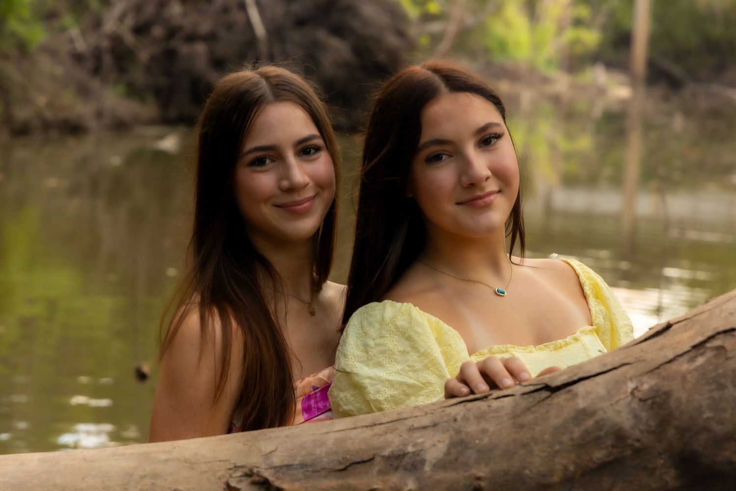 Two young women with long brown hair smiling together outdoors near a river, one wearing a yellow dress.