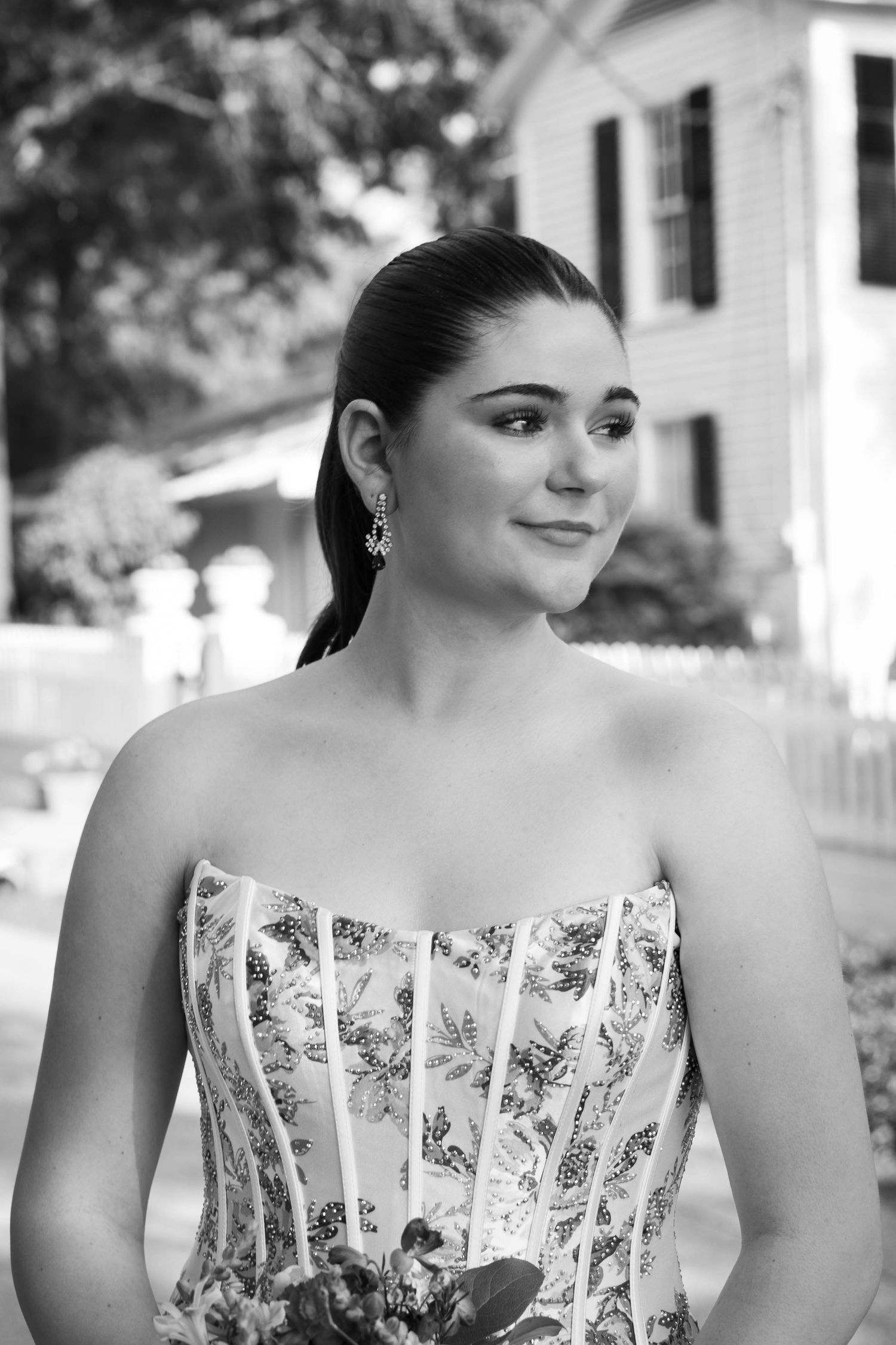 Black and white portrait of a young woman in a floral corset dress with elegant earrings, posing outdoors near a historic home.