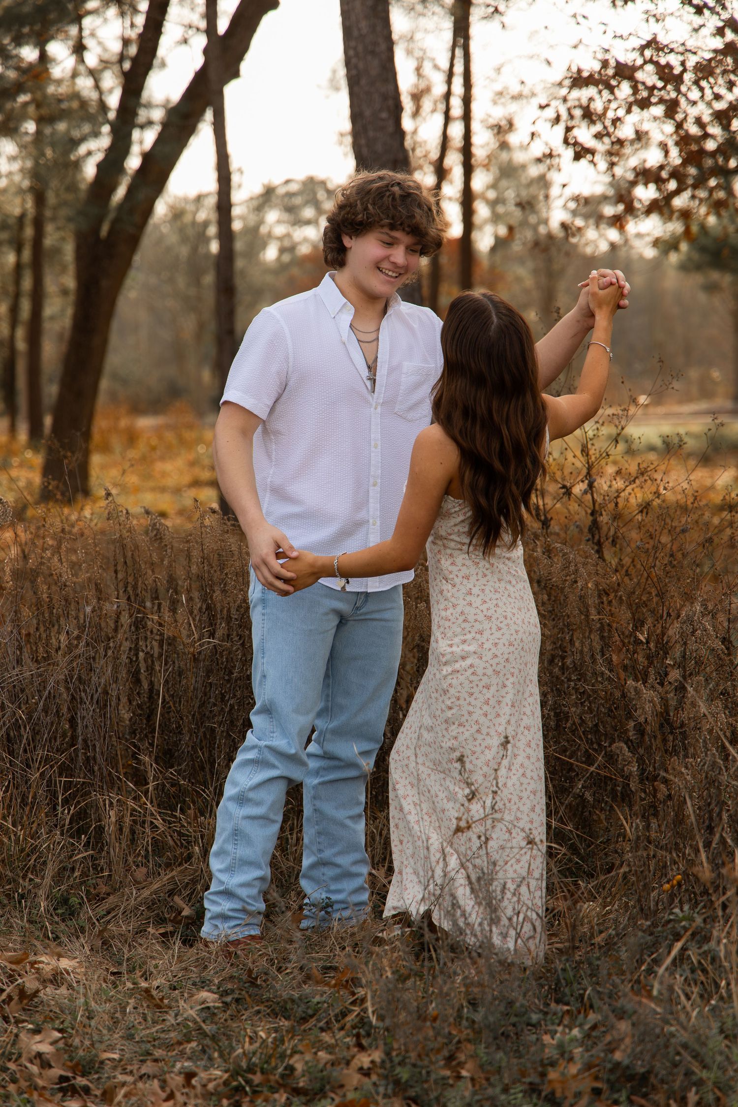 Young couple dancing together in autumn forest, man in white shirt and jeans smiling at woman in lace dress.