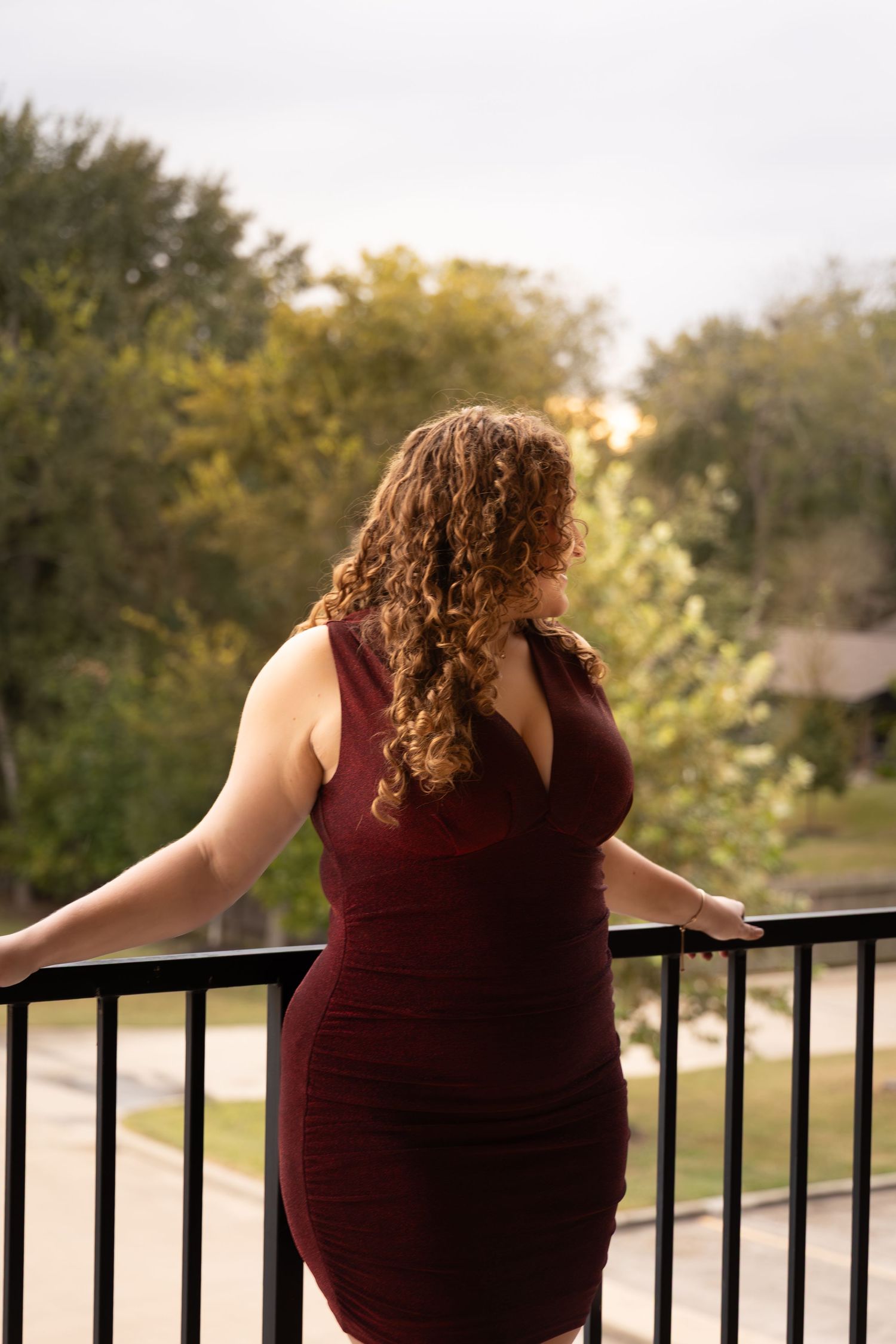 Woman in burgundy dress with curly hair stands on balcony overlooking green trees and suburban street.
