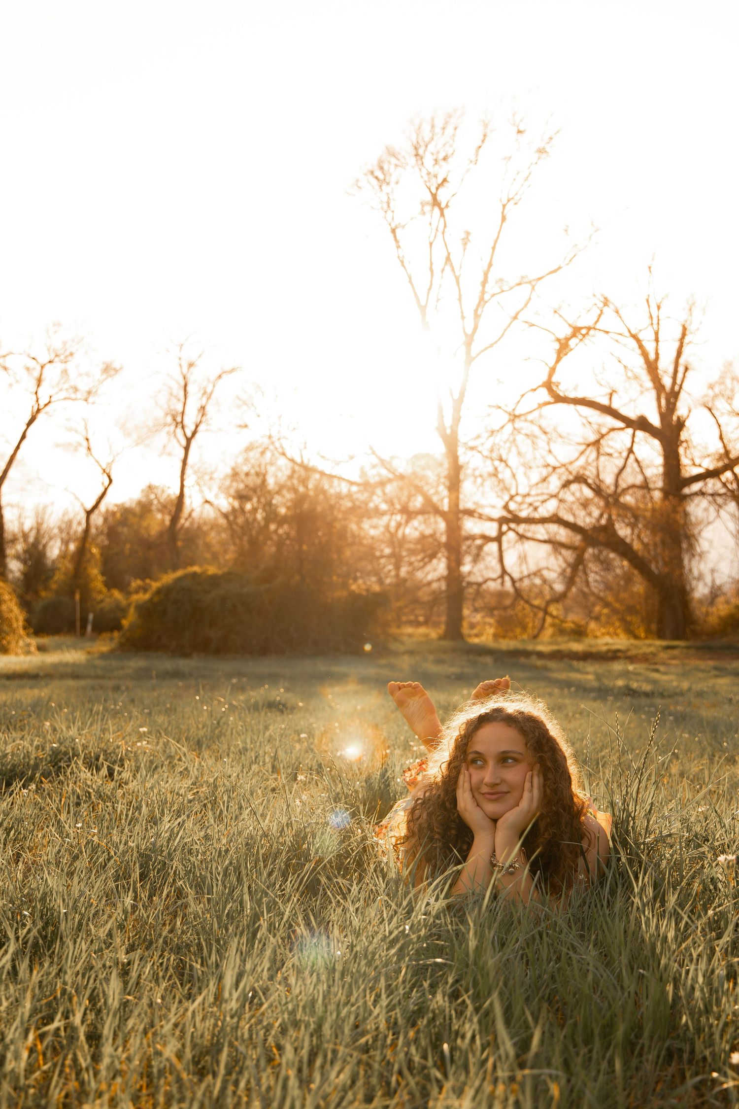 Young woman with curly hair lying in golden grass at sunset, smiling dreamily with bare trees glowing in warm backlight.
