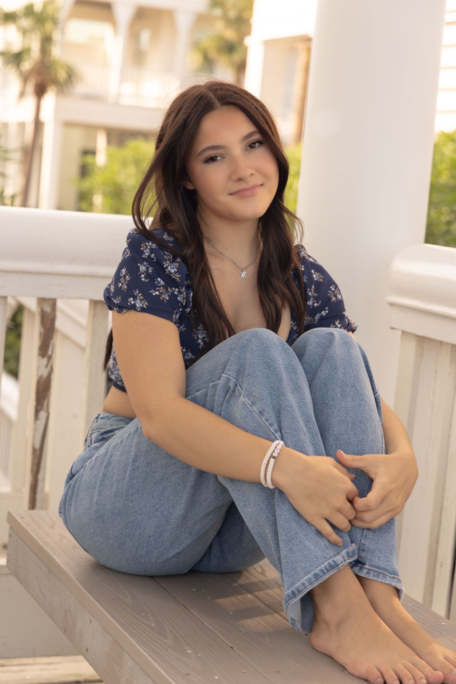 Smiling brunette teen girl sitting on wooden steps in jeans and navy floral top for senior photos.