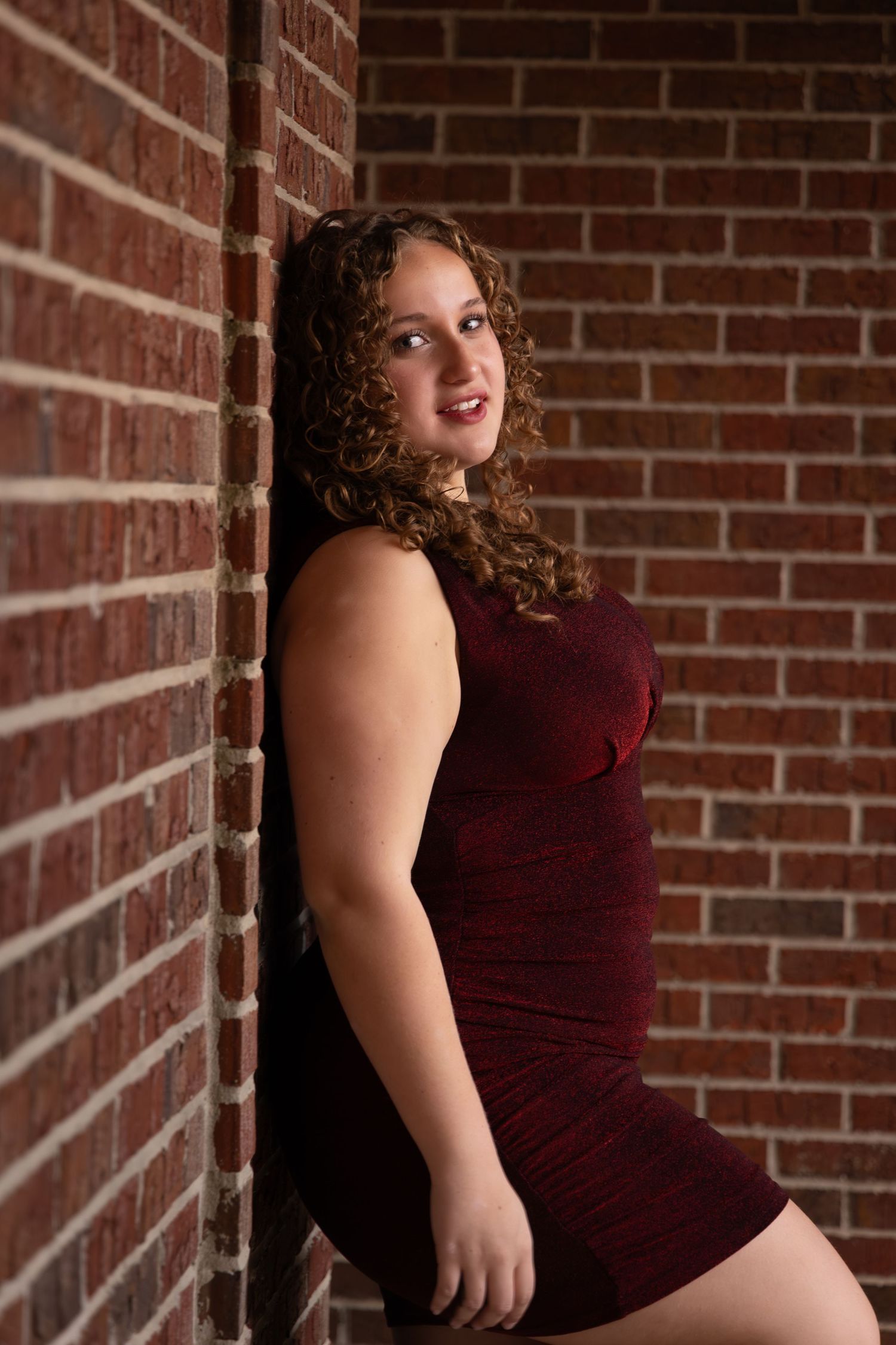 Young woman with curly hair in a burgundy dress leaning against a red brick wall, smiling confidently.