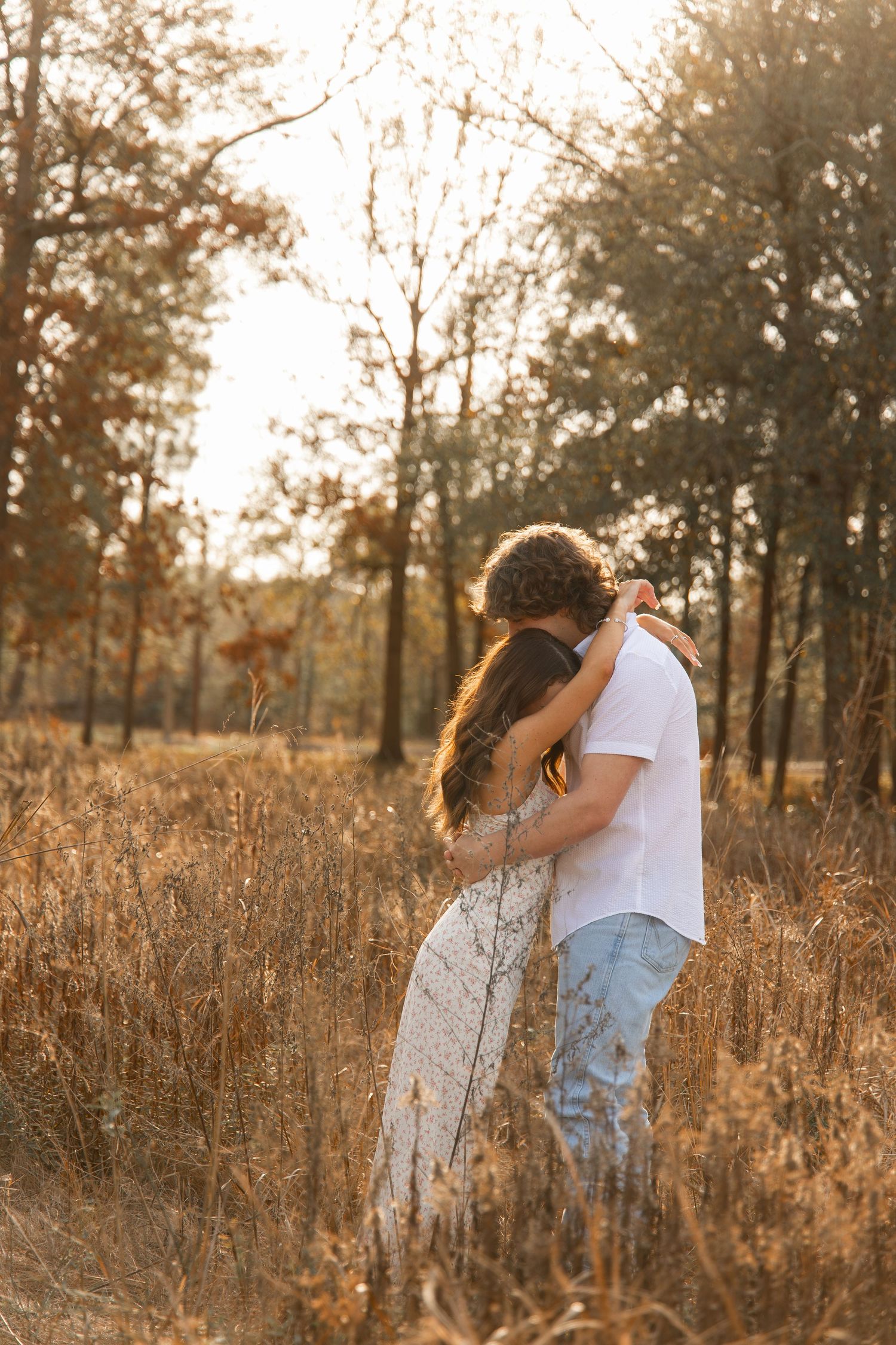 Romantic couple embracing in golden field at sunset, woman in white lace dress hugging man in white shirt.