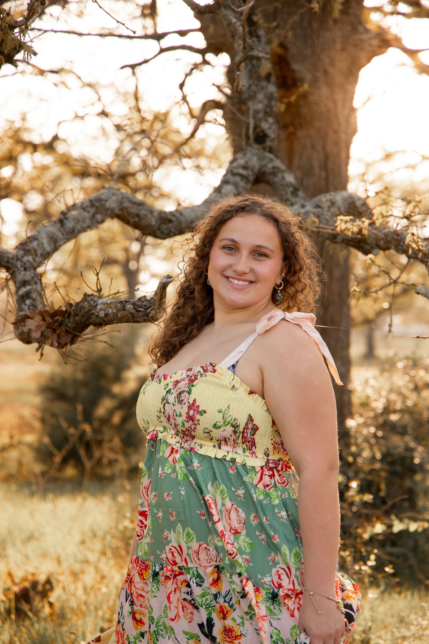 Young woman in floral maxi dress smiling outdoors by a large oak tree in golden sunset light.