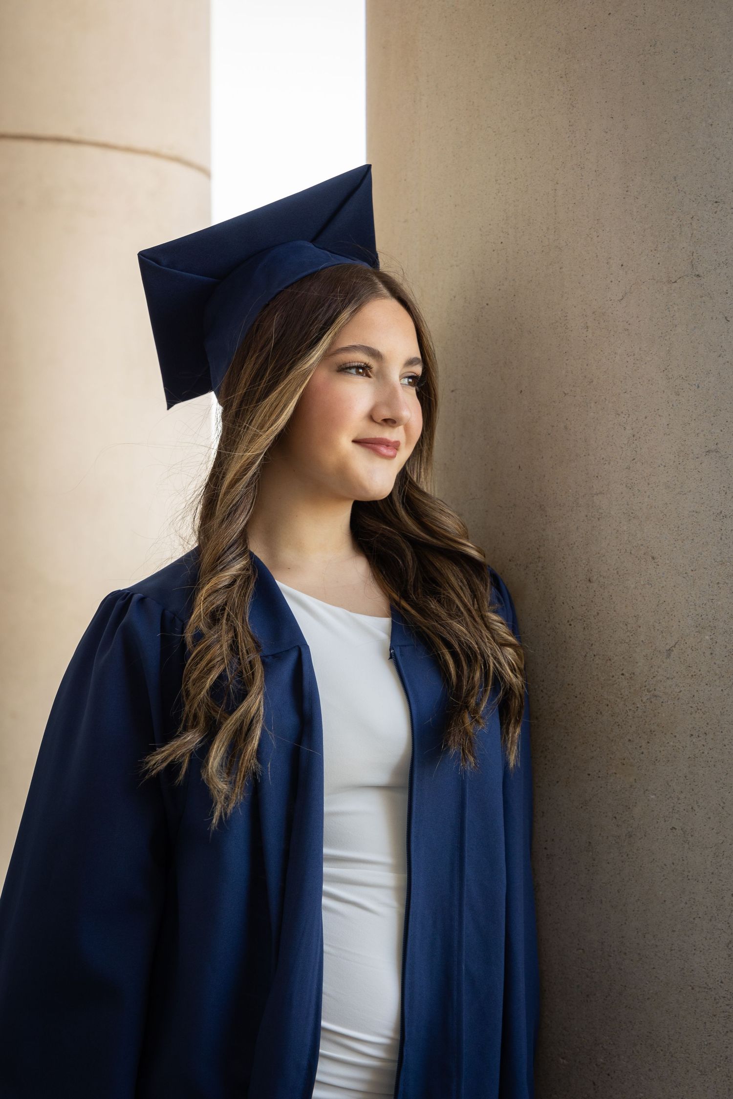 Graduate in navy blue cap and gown poses thoughtfully by stone pillars in natural light.
