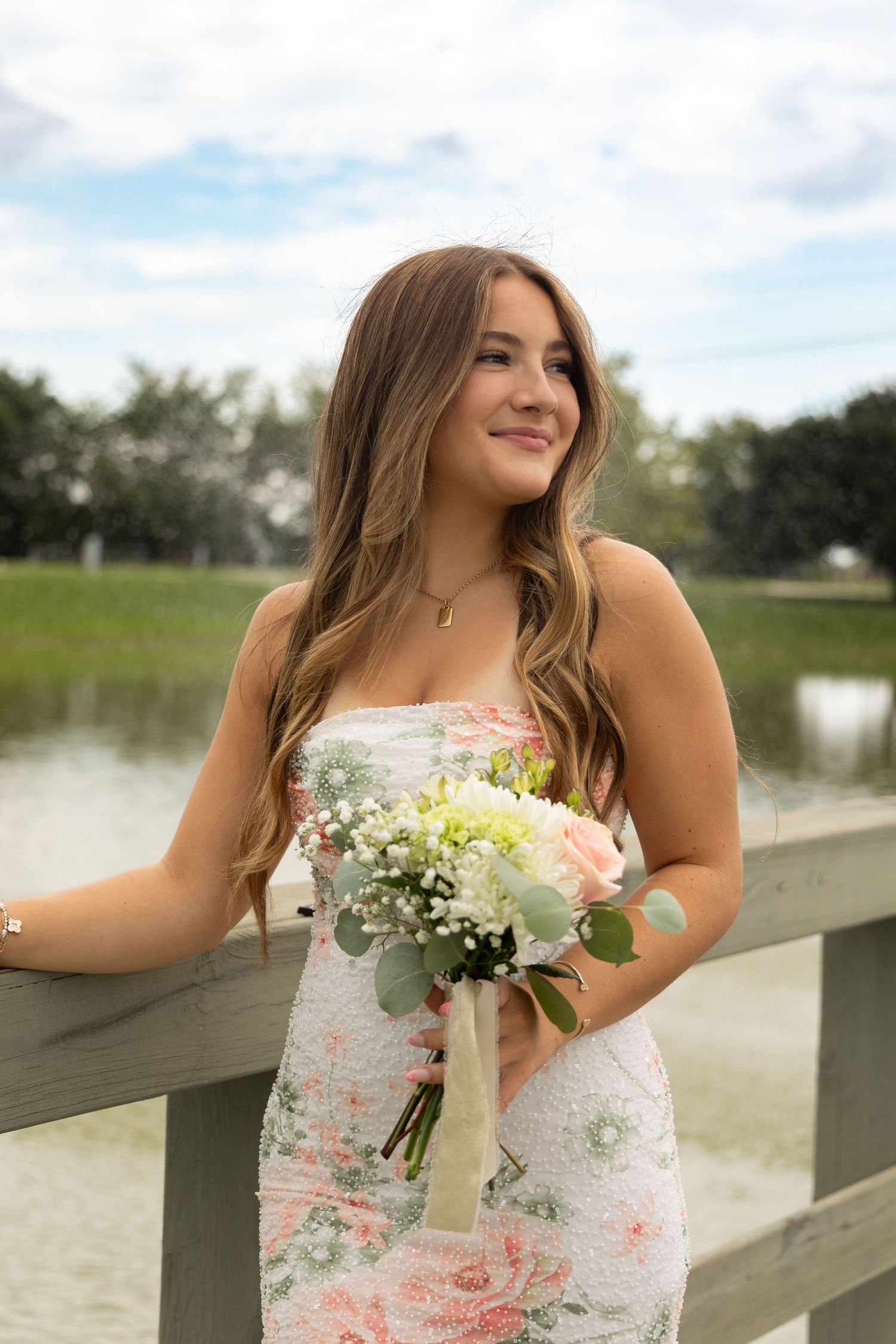 Young woman in floral strapless dress holding a bouquet of white and pink flowers on a wooden bridge.