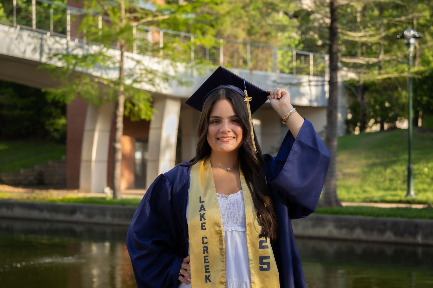 Smiling graduate in blue cap and gown with gold honor stole poses outdoors near a pond on graduation day.