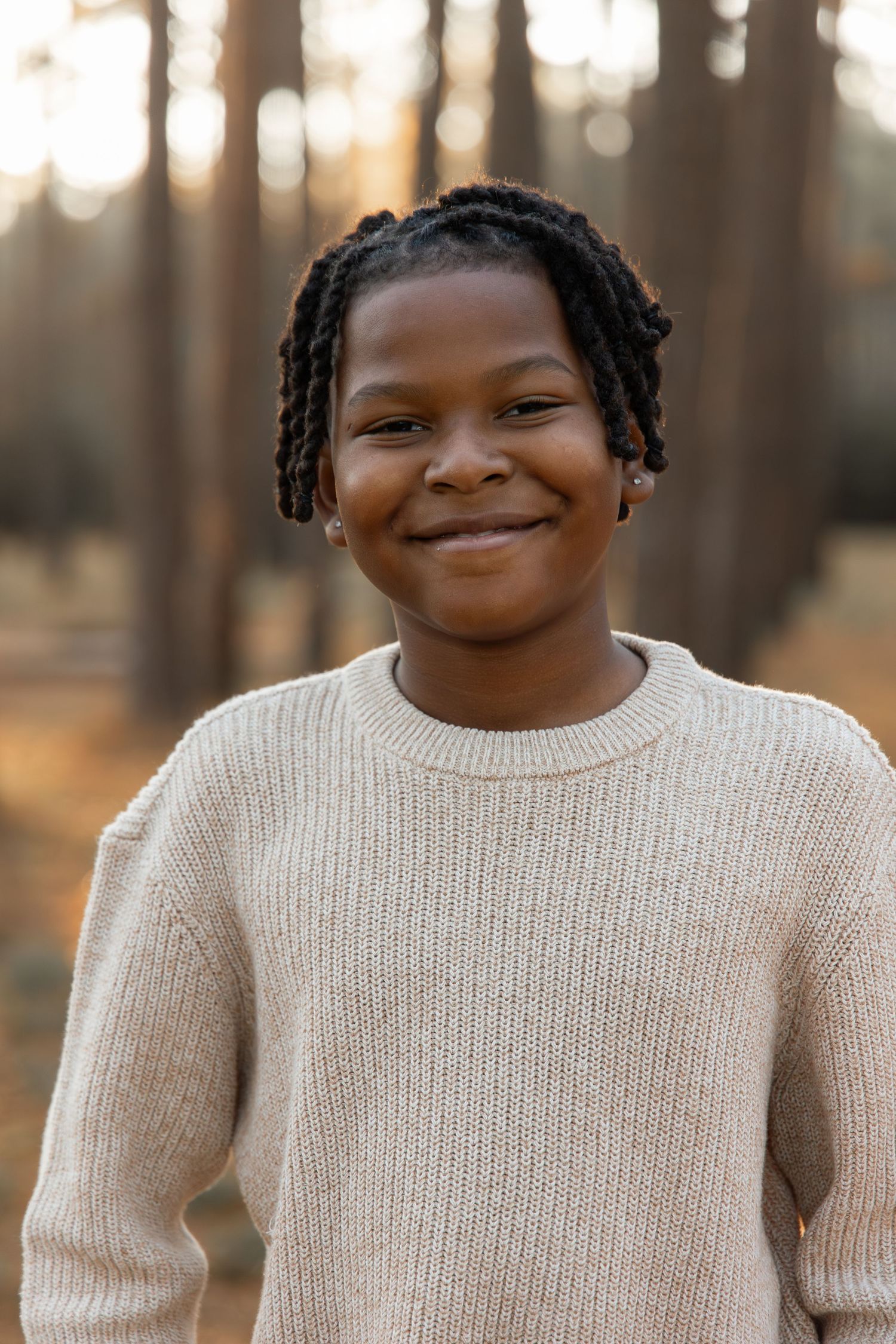 Smiling young Black boy with dreadlocks wearing a beige knit sweater, standing outdoors in a pine forest.