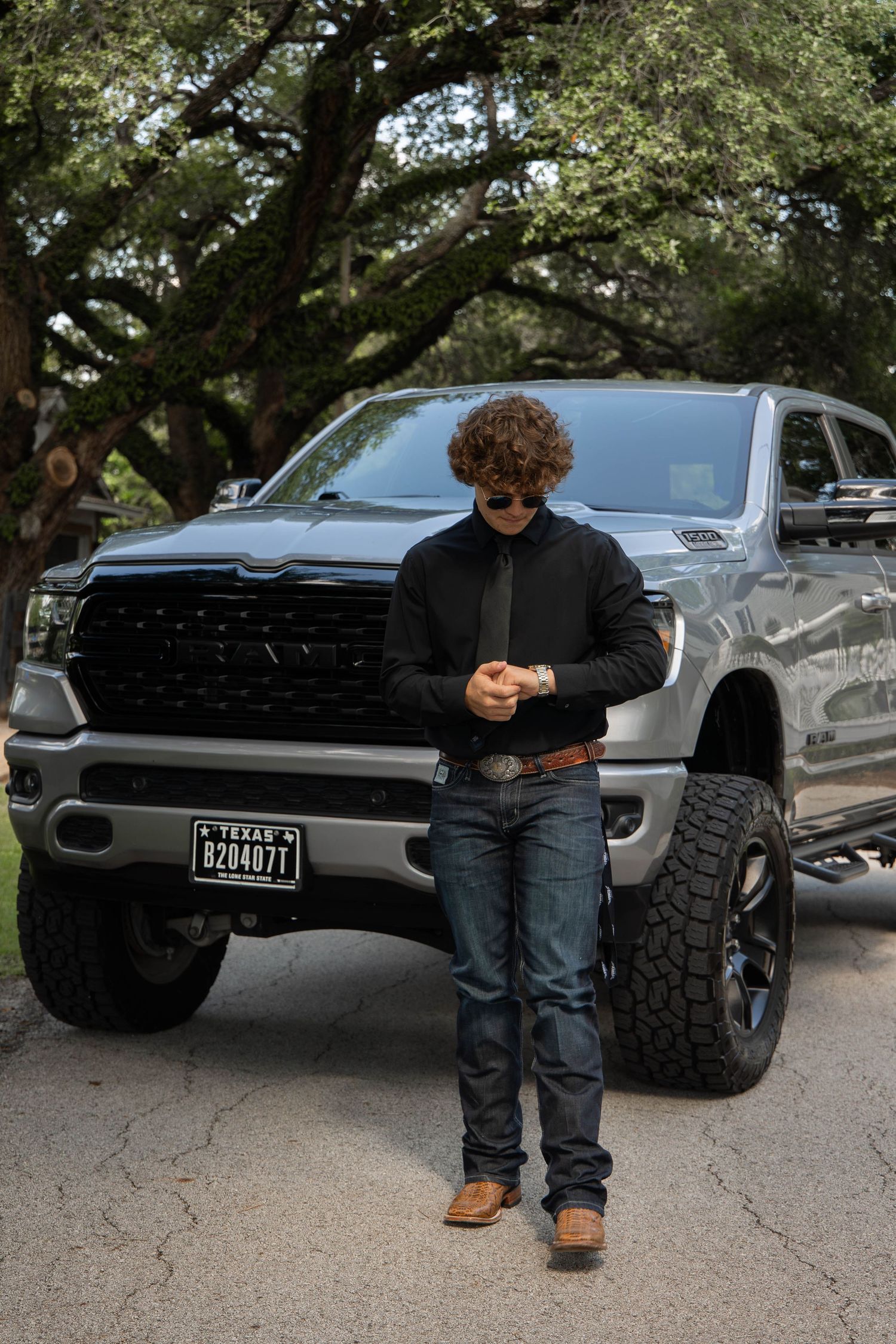Young man with curly hair in black jacket and jeans stands before a lifted gray Ram 1500 truck with Texas plates.