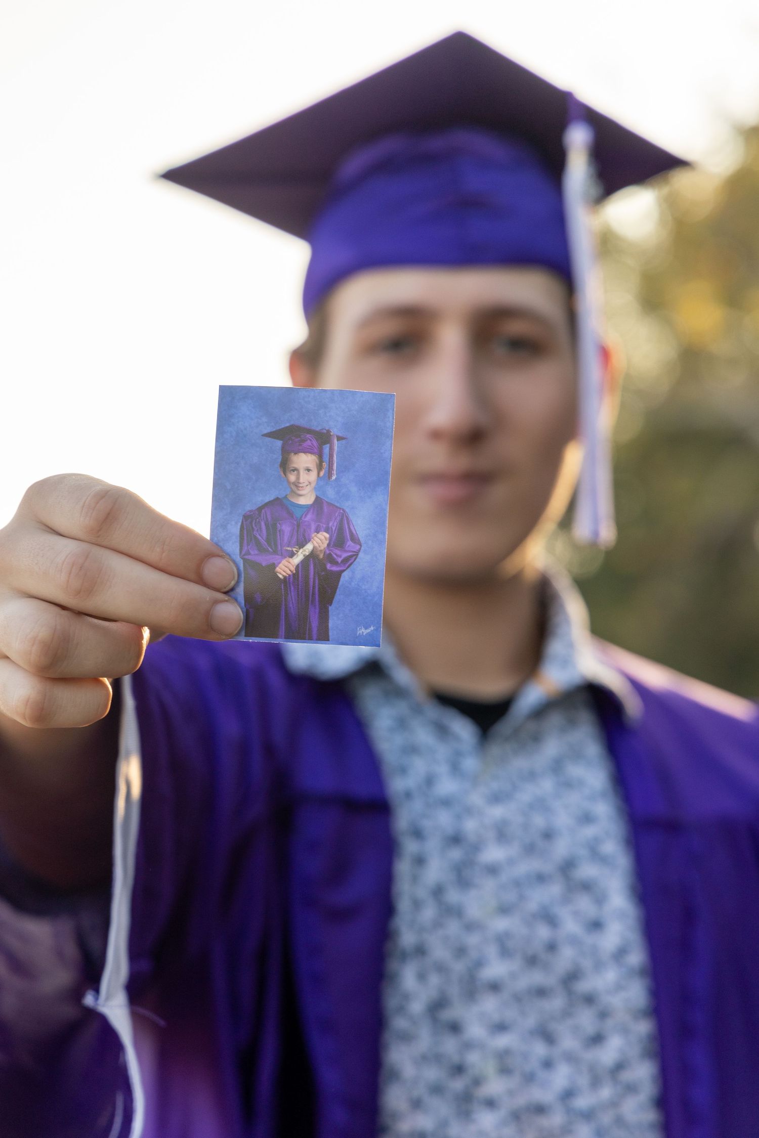 Graduate in purple cap and gown holds up a small portrait photo while standing outdoors.