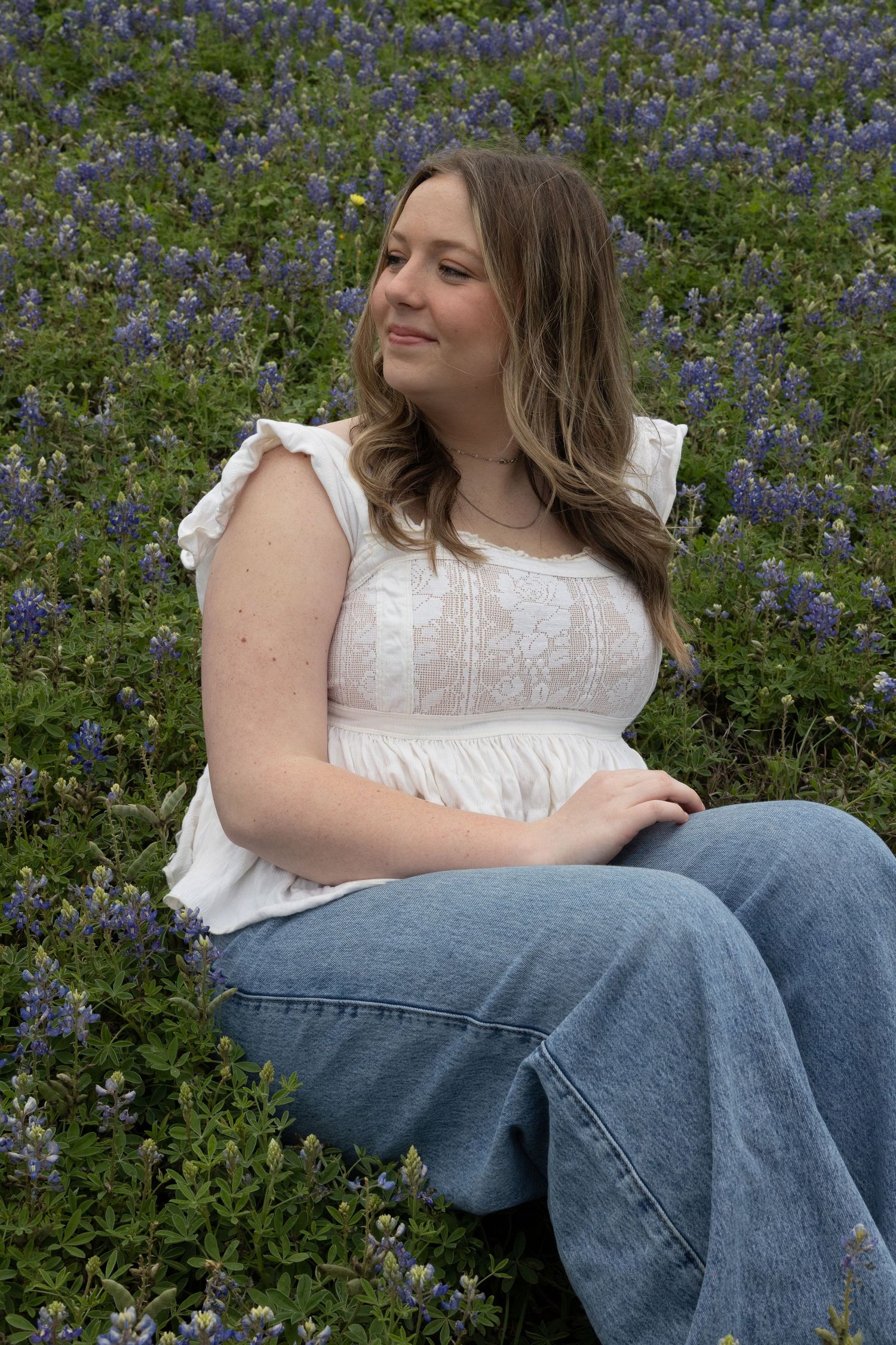 A person in a white top sits among blooming bluebonnets wearing blue jeans in an outdoor setting.