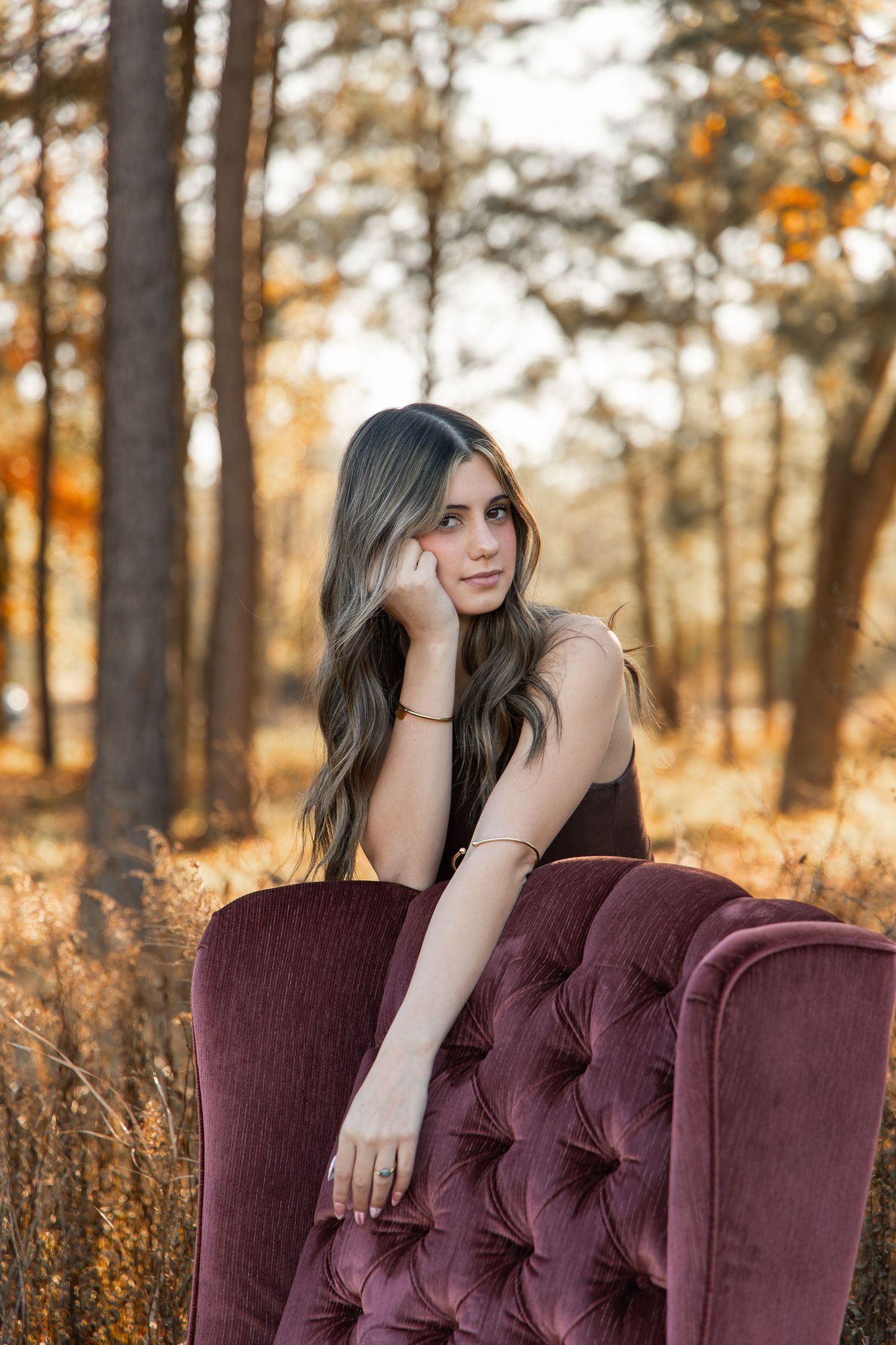 Young woman with long wavy hair poses on a velvet tufted chair outdoors in an autumn forest setting.