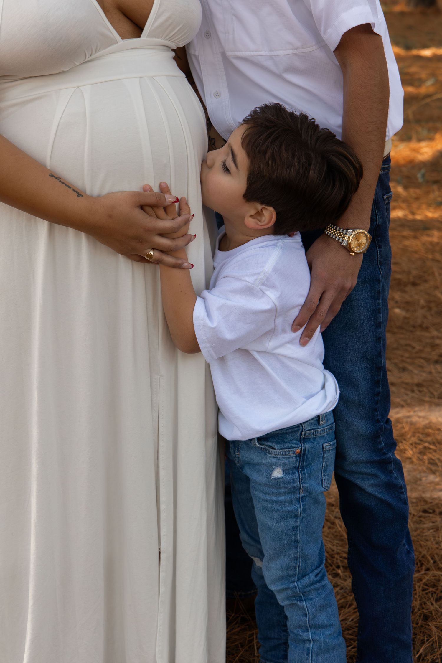A young boy kisses his mother's pregnant belly while standing between his parents outdoors.