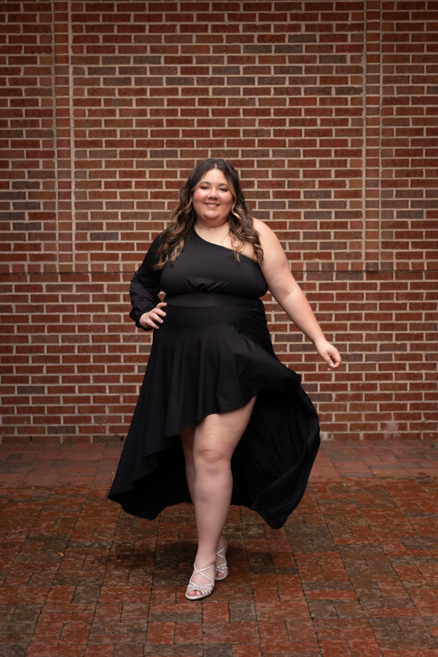 Woman in black asymmetric dress posing confidently in front of a red brick wall on a brick paved floor.