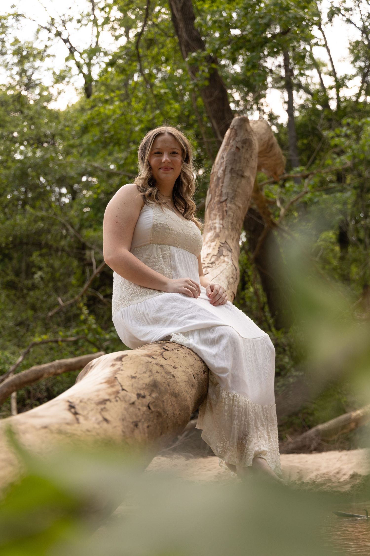 Woman in white dress sitting on fallen log in lush green forest setting.