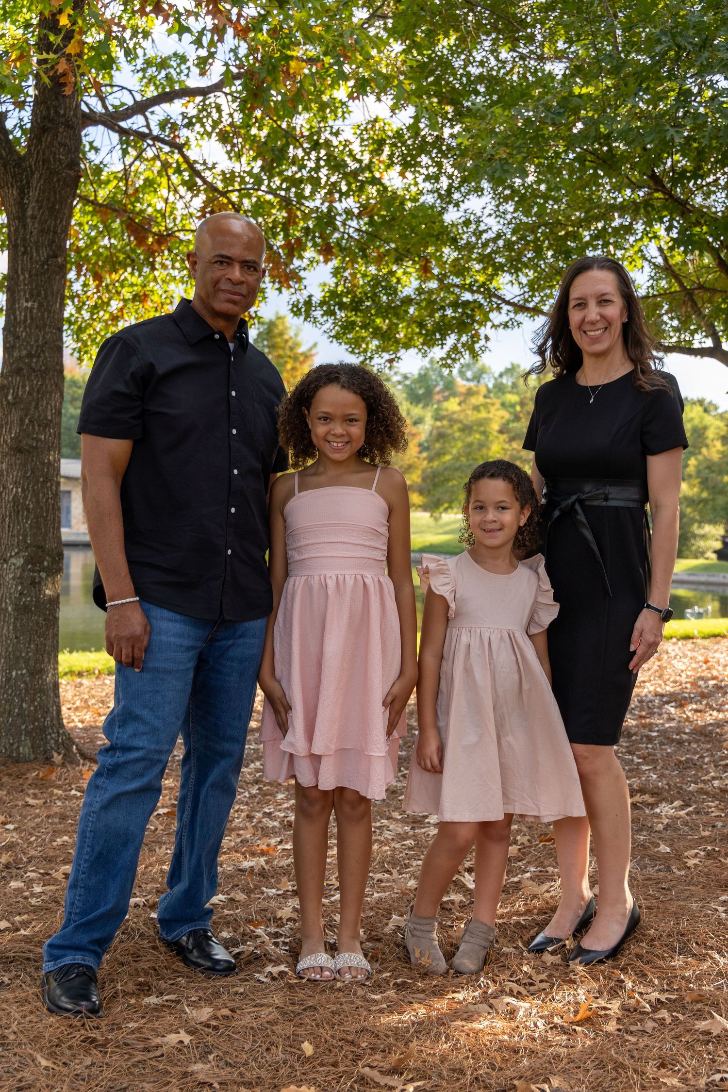 Family of four posing outdoors under autumn trees, parents with two young daughters in pink dresses.