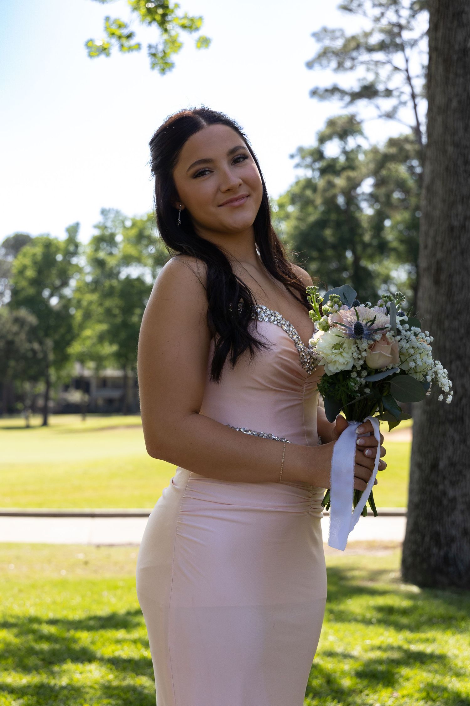 Young woman in pink gown holding white floral bouquet poses outdoors in park setting on sunny day.
