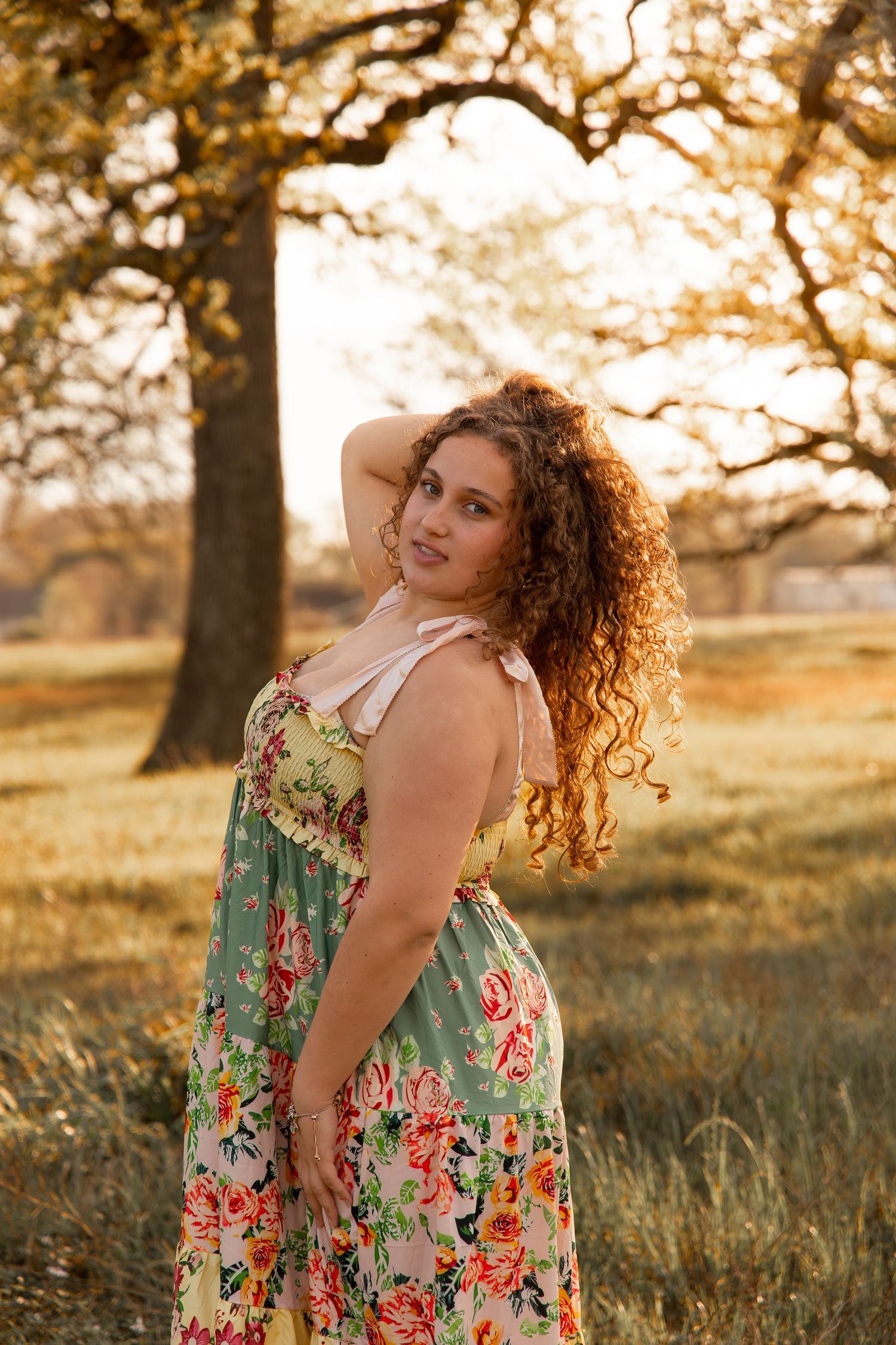 Curly-haired woman in floral maxi dress posing outdoors during golden hour with trees in background.
