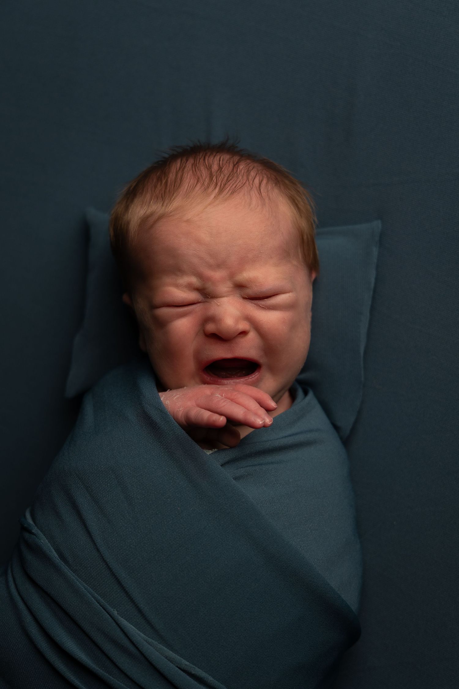Newborn baby swaddled in teal wrap crying on matching teal background during professional newborn photo session.