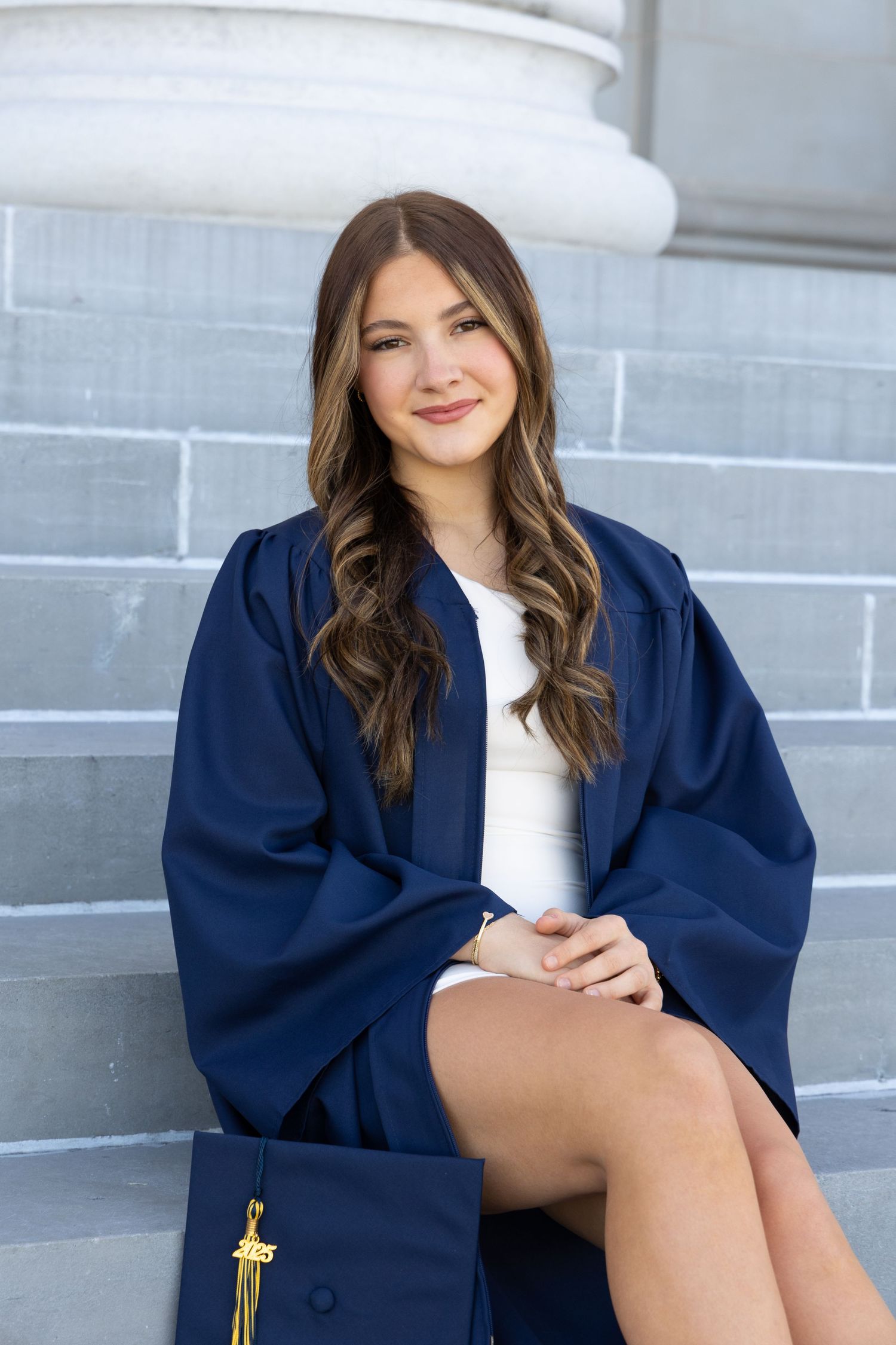 Young woman in navy graduation gown smiling on stone steps, holding cap with gold tassel.