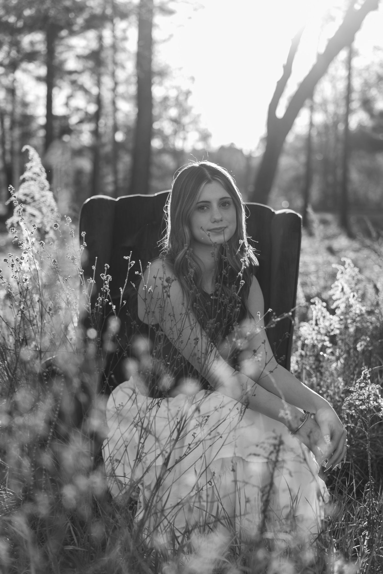 Black and white photo of a young woman in a floral dress sitting in a chair surrounded by wildflowers in a forest.