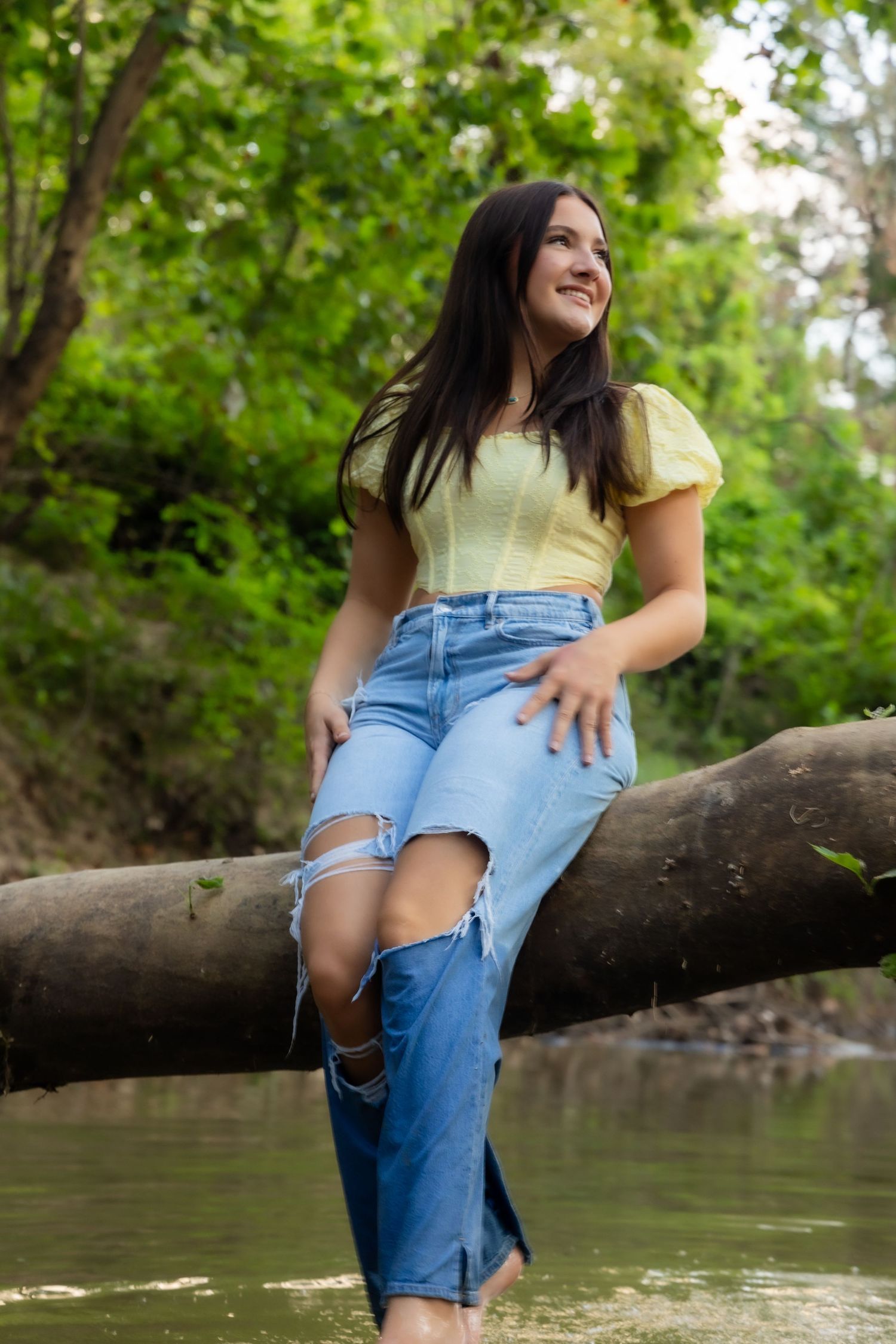 Young woman in yellow top and ripped jeans sitting on a log over a creek, smiling in a lush green forest.