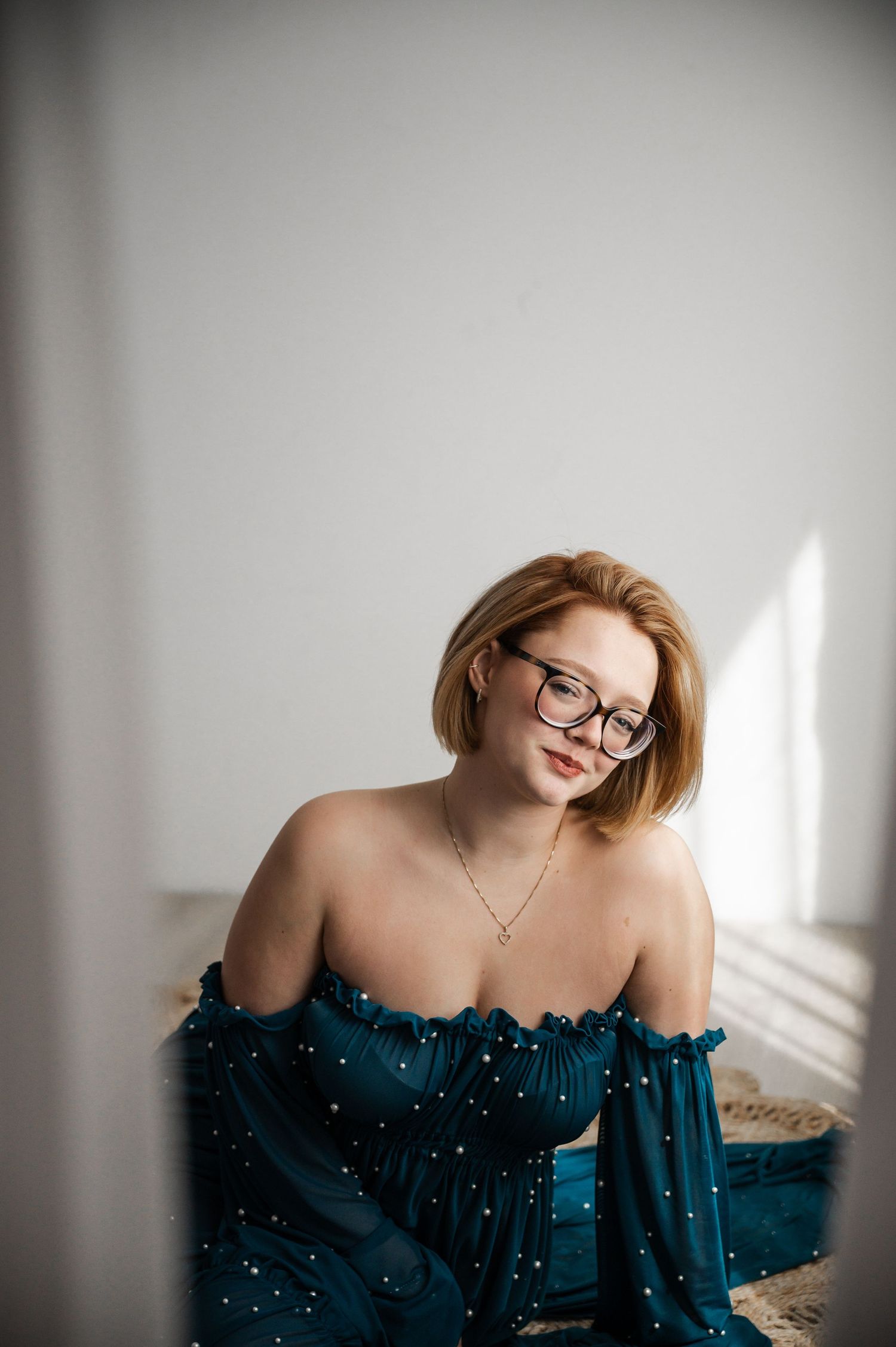 Young woman in teal off-shoulder gown with pearl details, wearing glasses and gold necklace, seated against white wall.