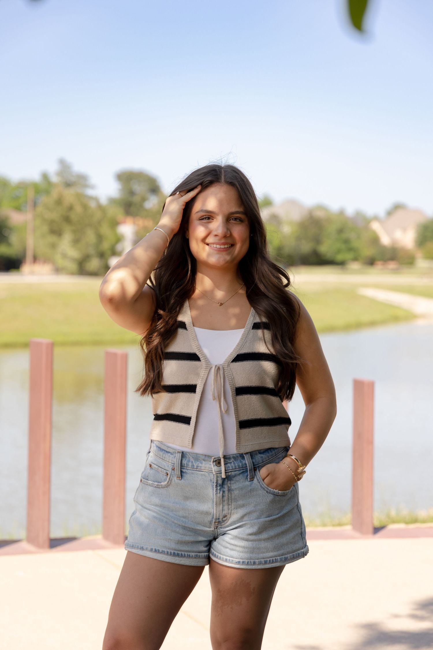 Young woman in striped vest and denim shorts posing outdoors near a lake on a sunny day.