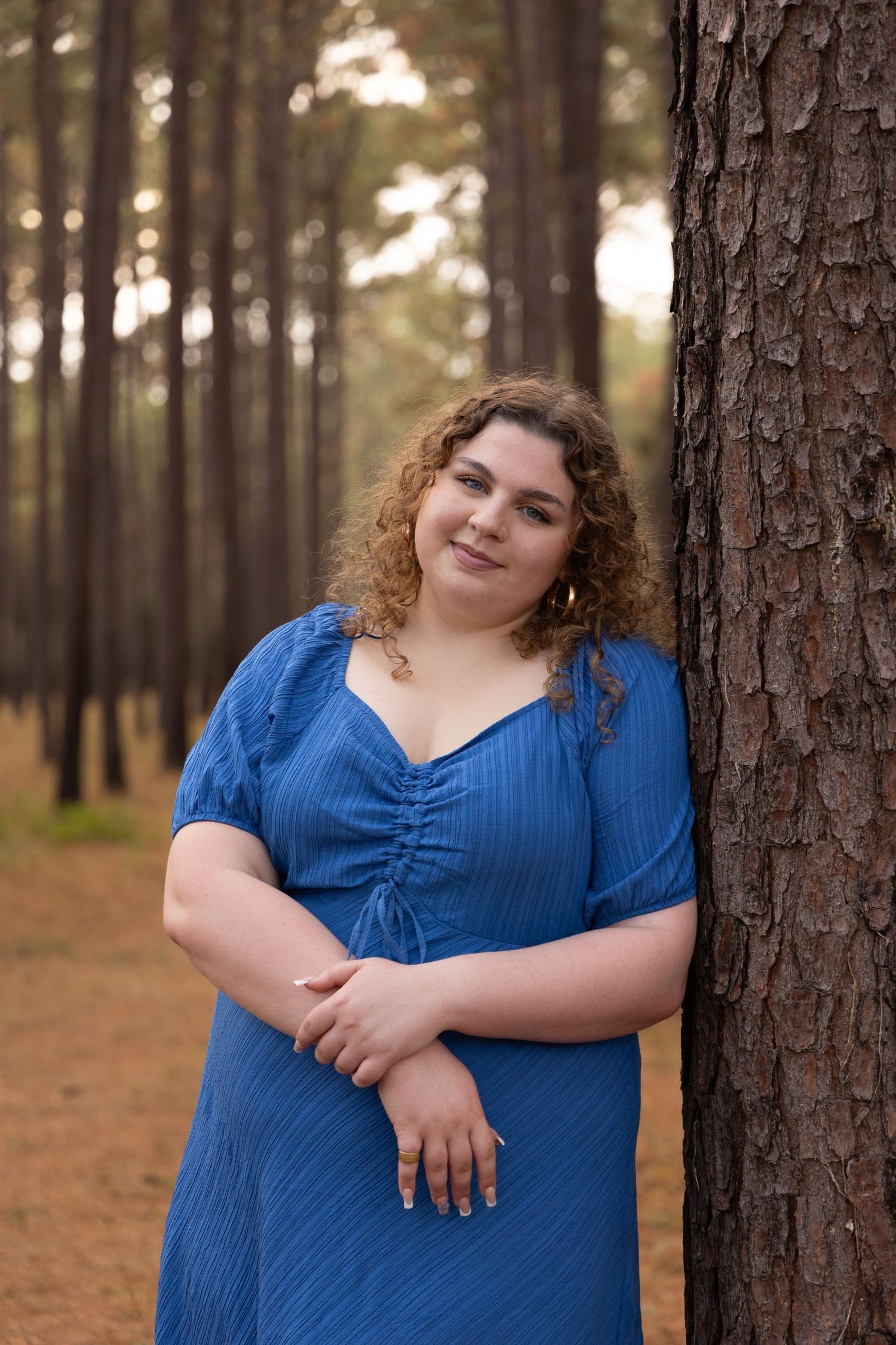 A curly-haired woman in a blue dress leans against a pine tree in a forest, smiling warmly at the camera.