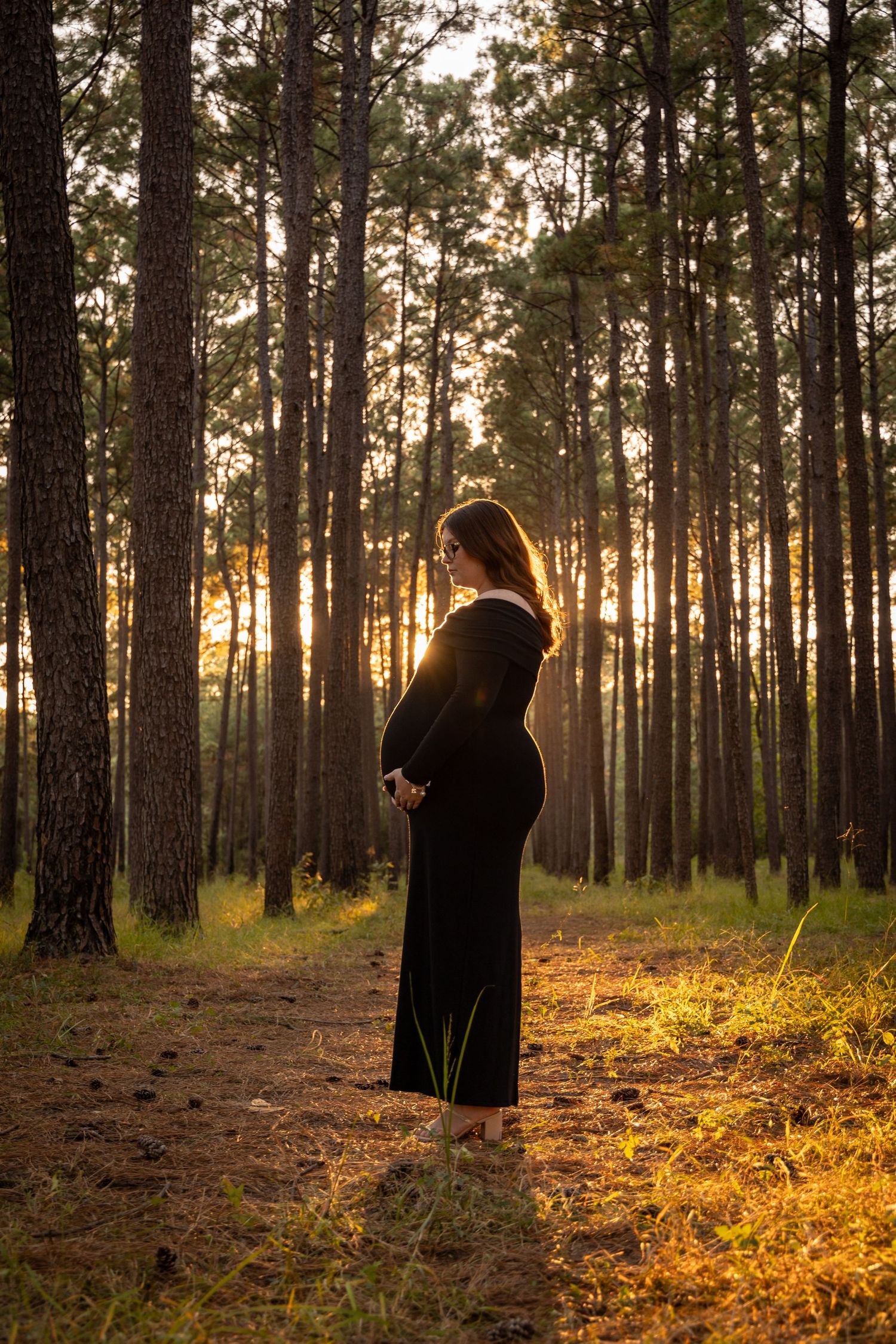 Expectant mother stands in profile against sunlit forest backdrop at golden hour.